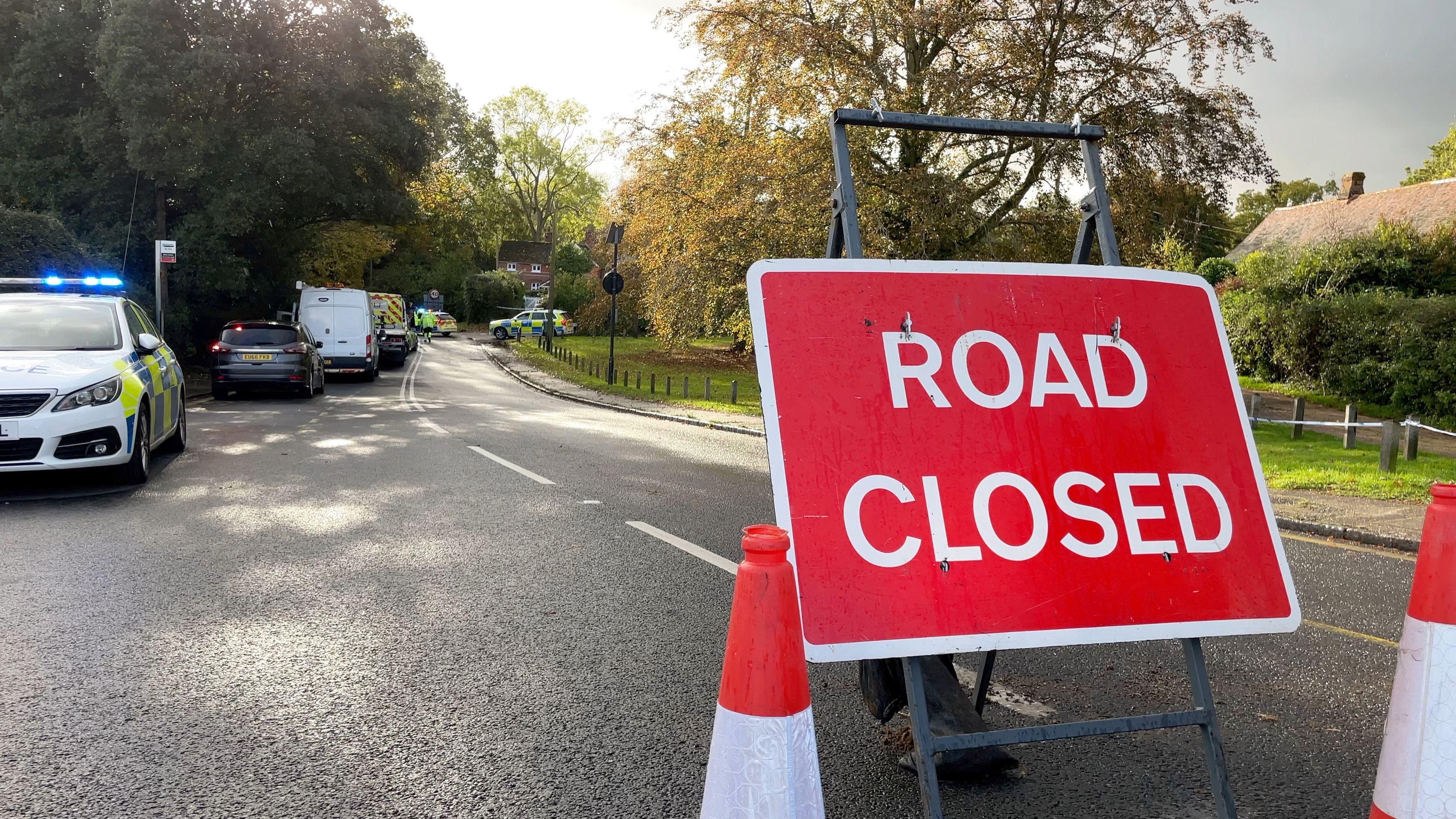 A road is closed off with orange cones and a large red 'road closed' sign on a stand. Further down the closed road are seven police vehicles. One has its blue emergency lights on.