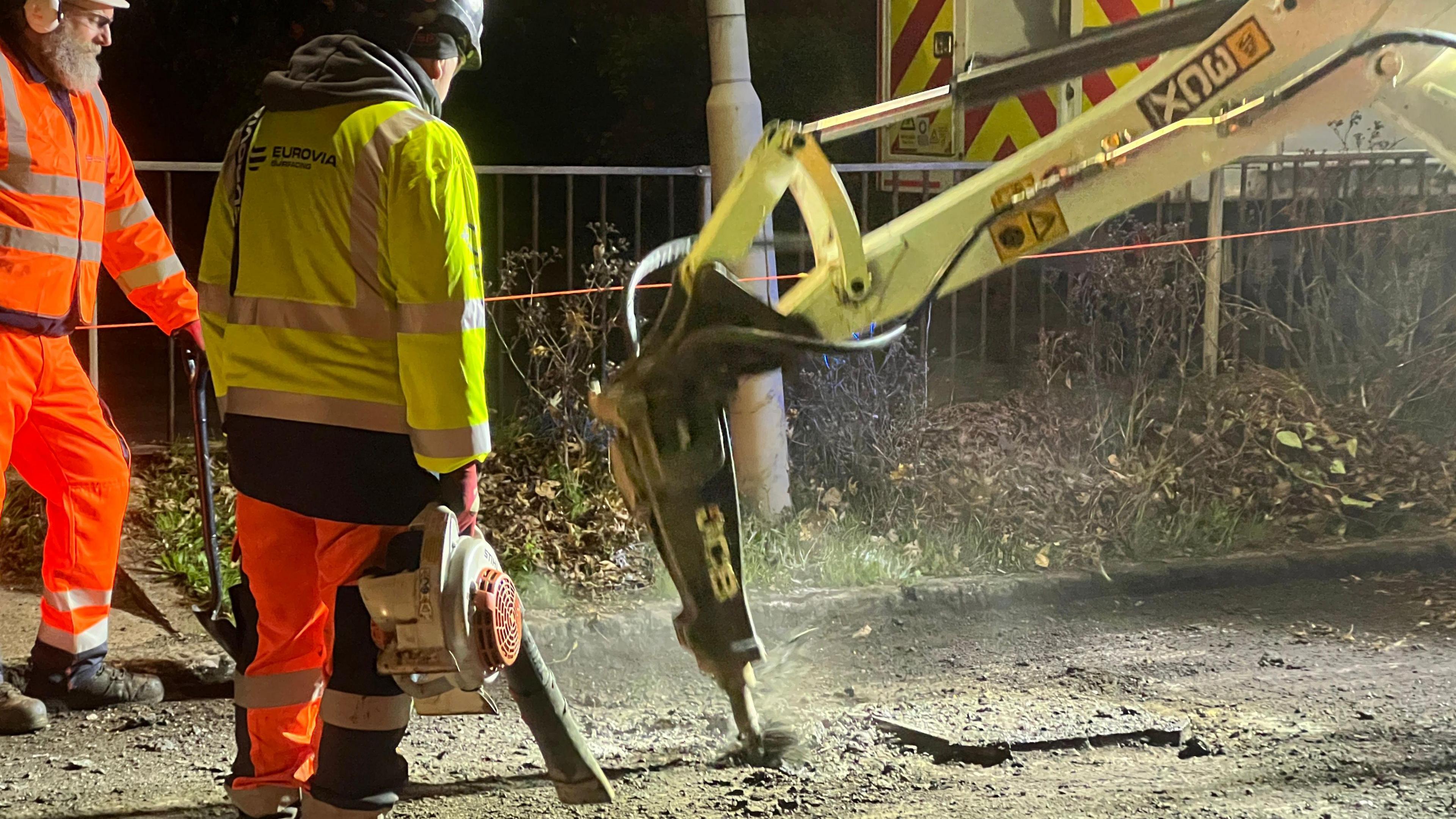 Two men wearing high-visibility jackets and helmets watch as heavy machinery digs up a road surface. It is dark.