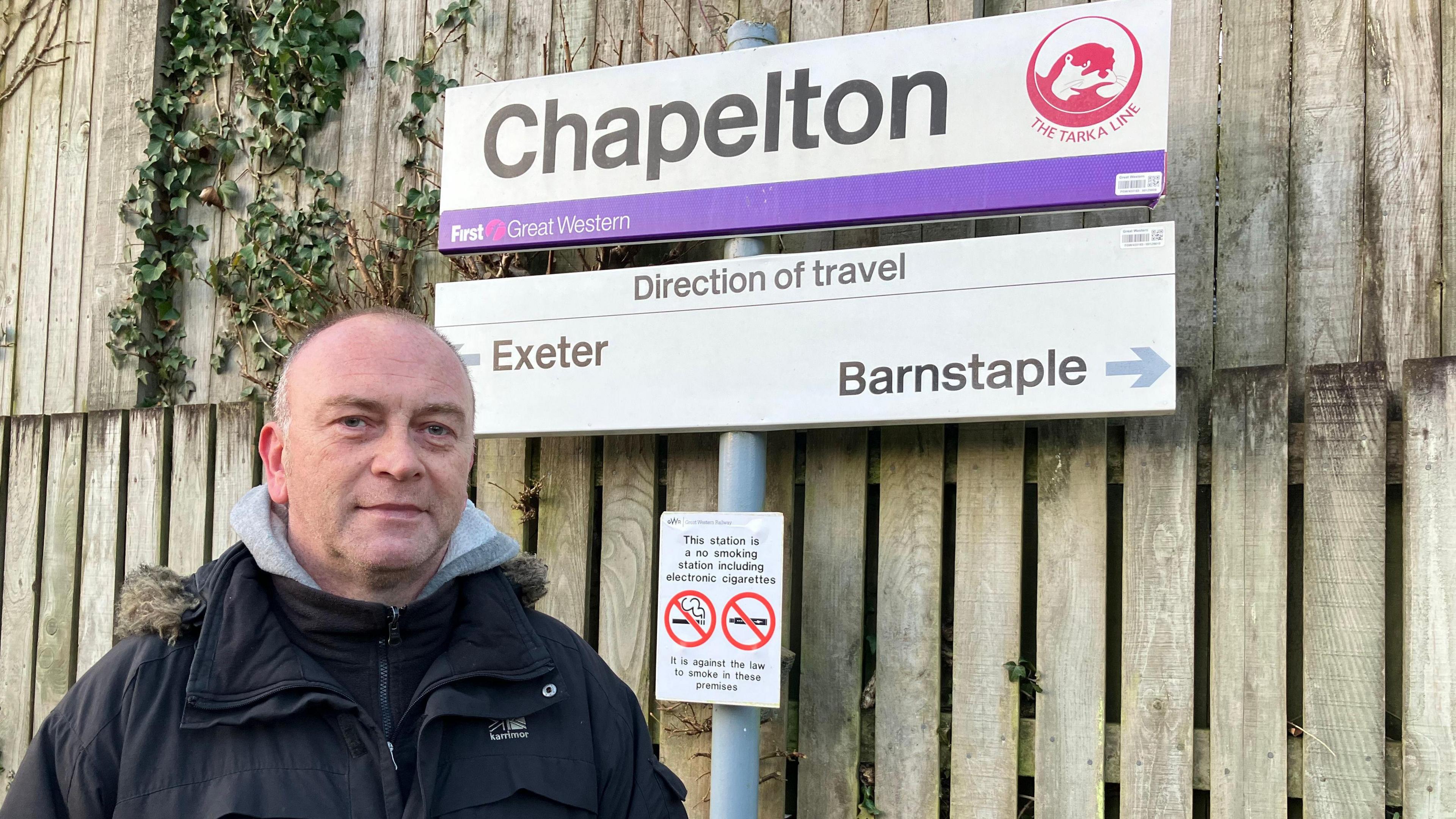 A  man with short hair and wearing a navy parker coat over a grey hoodie, He is standing in front of the Chapelton Train Station sign which is in front of a fence The signpost also includes a direction of travel sign pointing to Exeter on the left and Barnstaple on the right