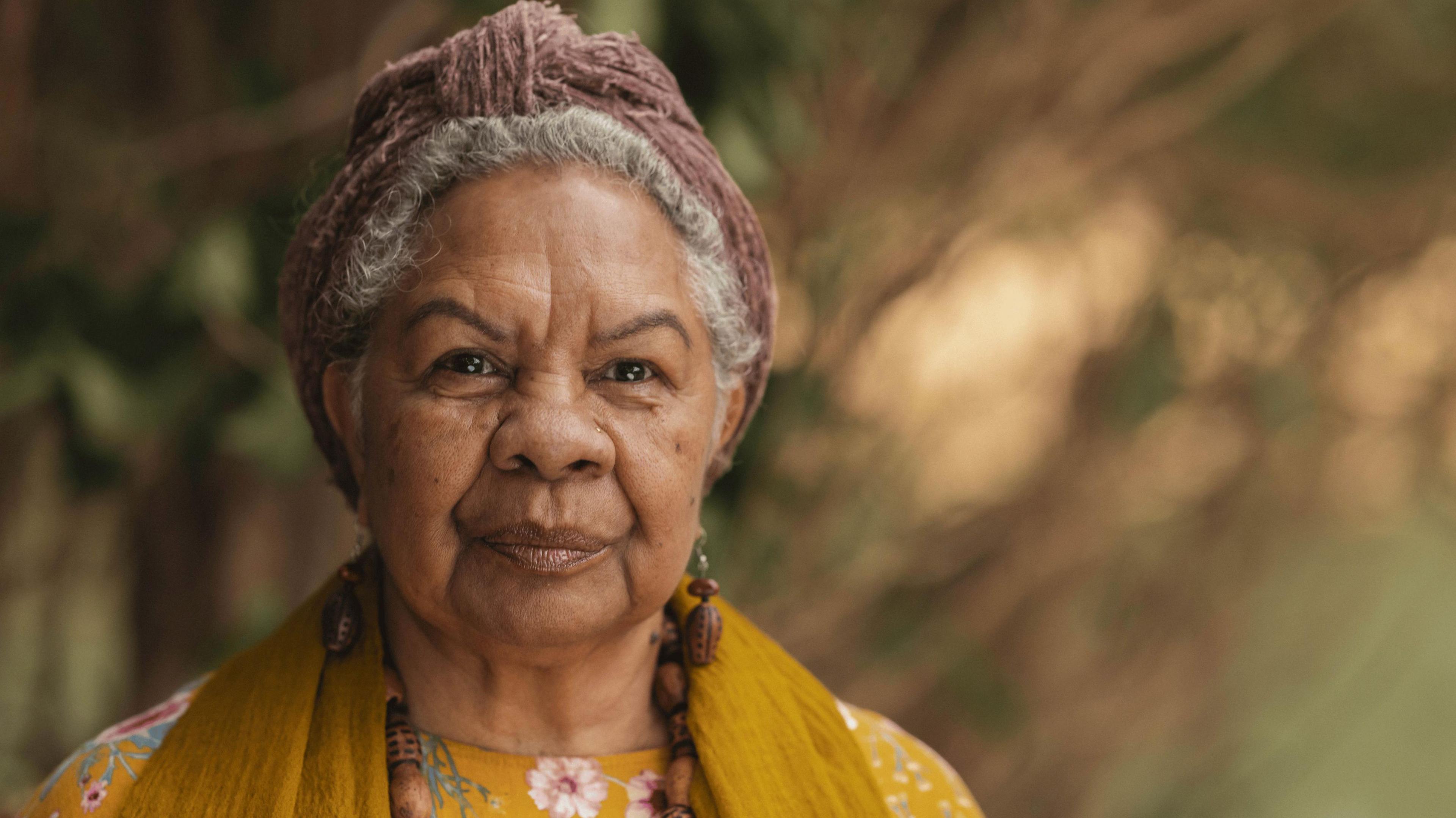 A woman with grey hair tied up with a light brown headband, wearing a yellow floral top and a mustard-coloured scarf