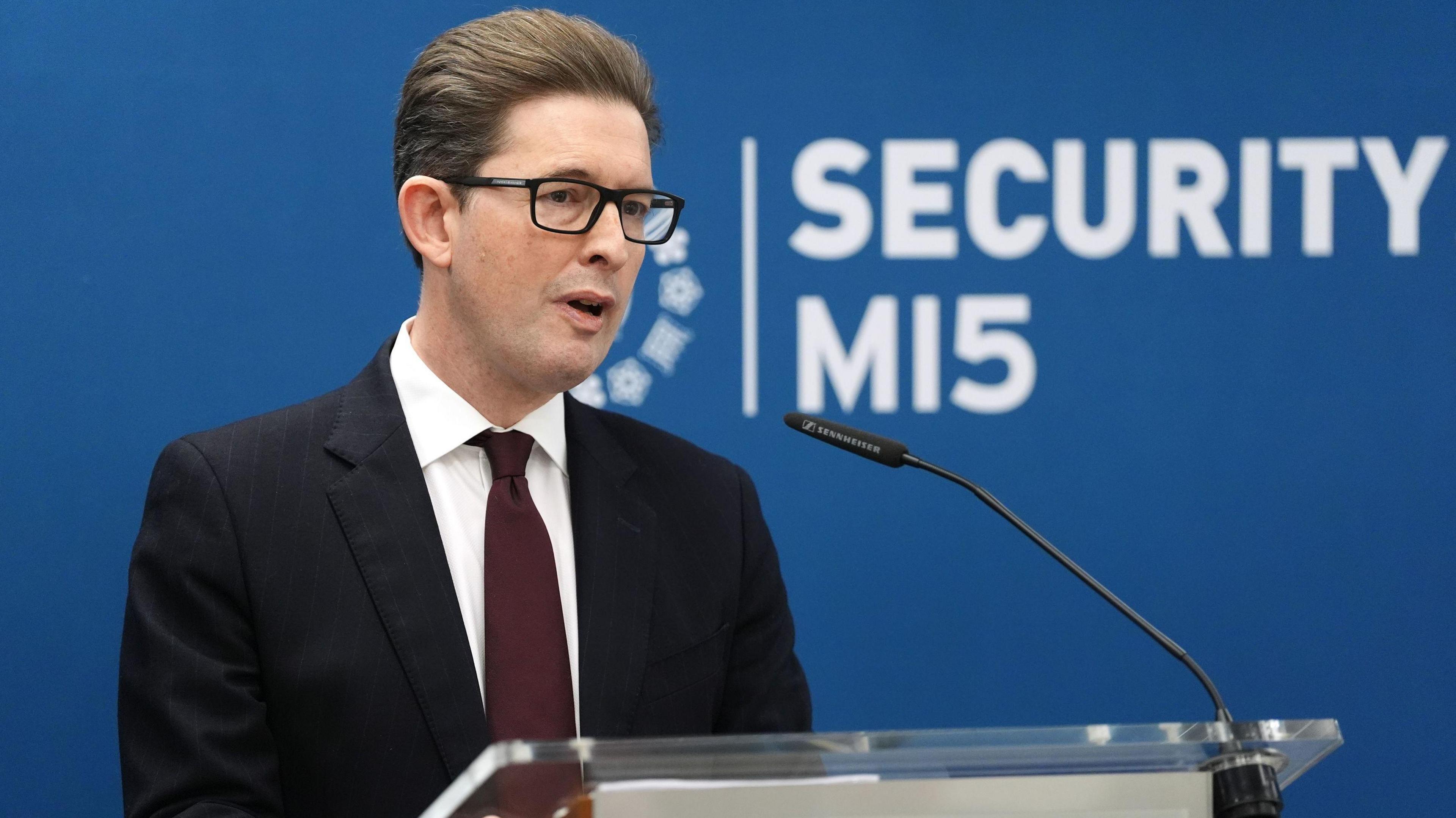 Ken McCallum, Director General of MI5 - a middle-aged antheral   with slicked backmost  acheronian  brownish  hair, and wearing dark-rimmed rectangular glasses. He is wearing a acheronian  suit, achromatic  garment  and navy bluish  tie.