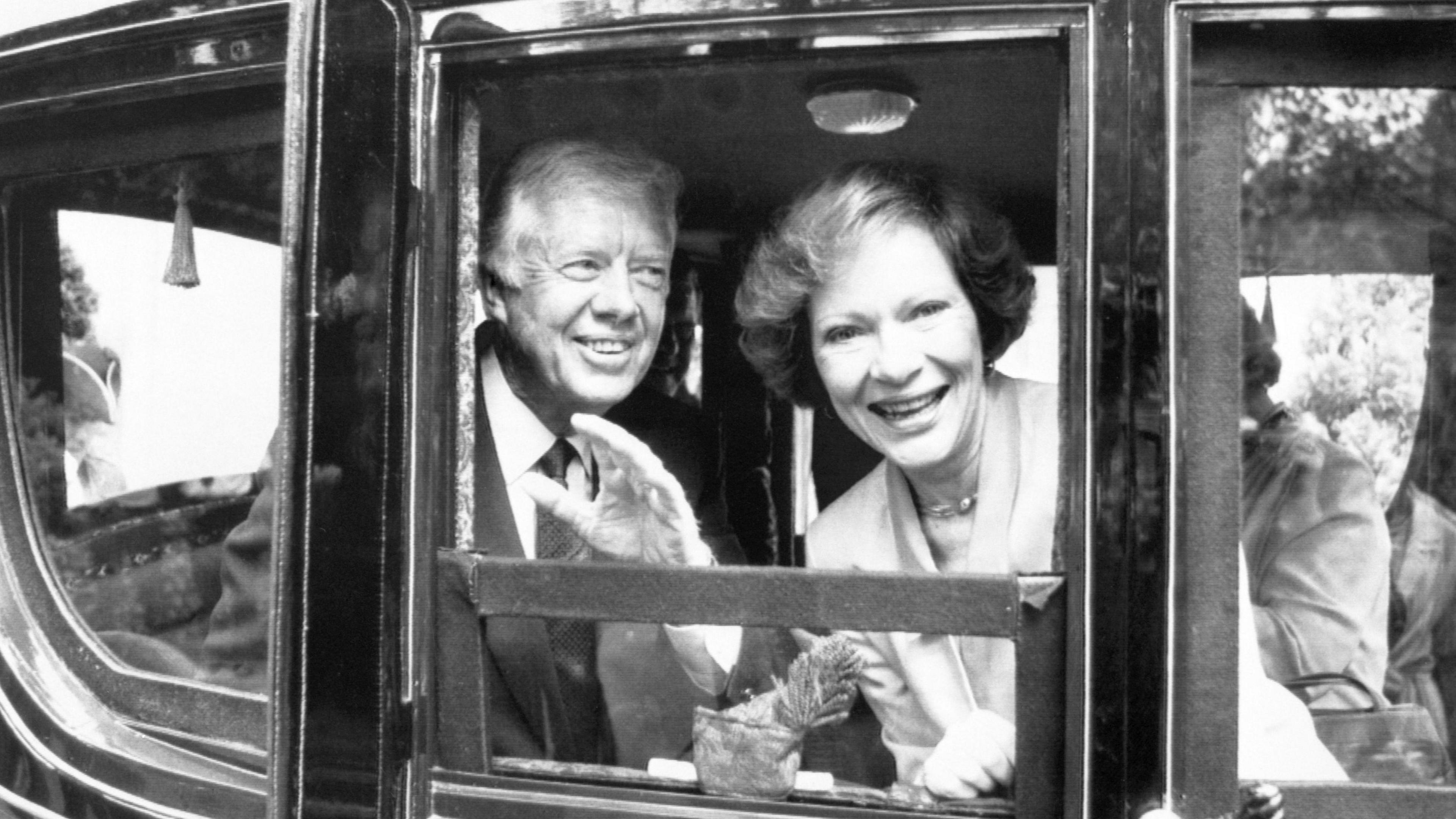 A black and white photo of Carter and his wife, Rosalynn, in a black carriage. Rosalynn is looking into the camera, smiling while Jimmy Carter looks to the side waving.