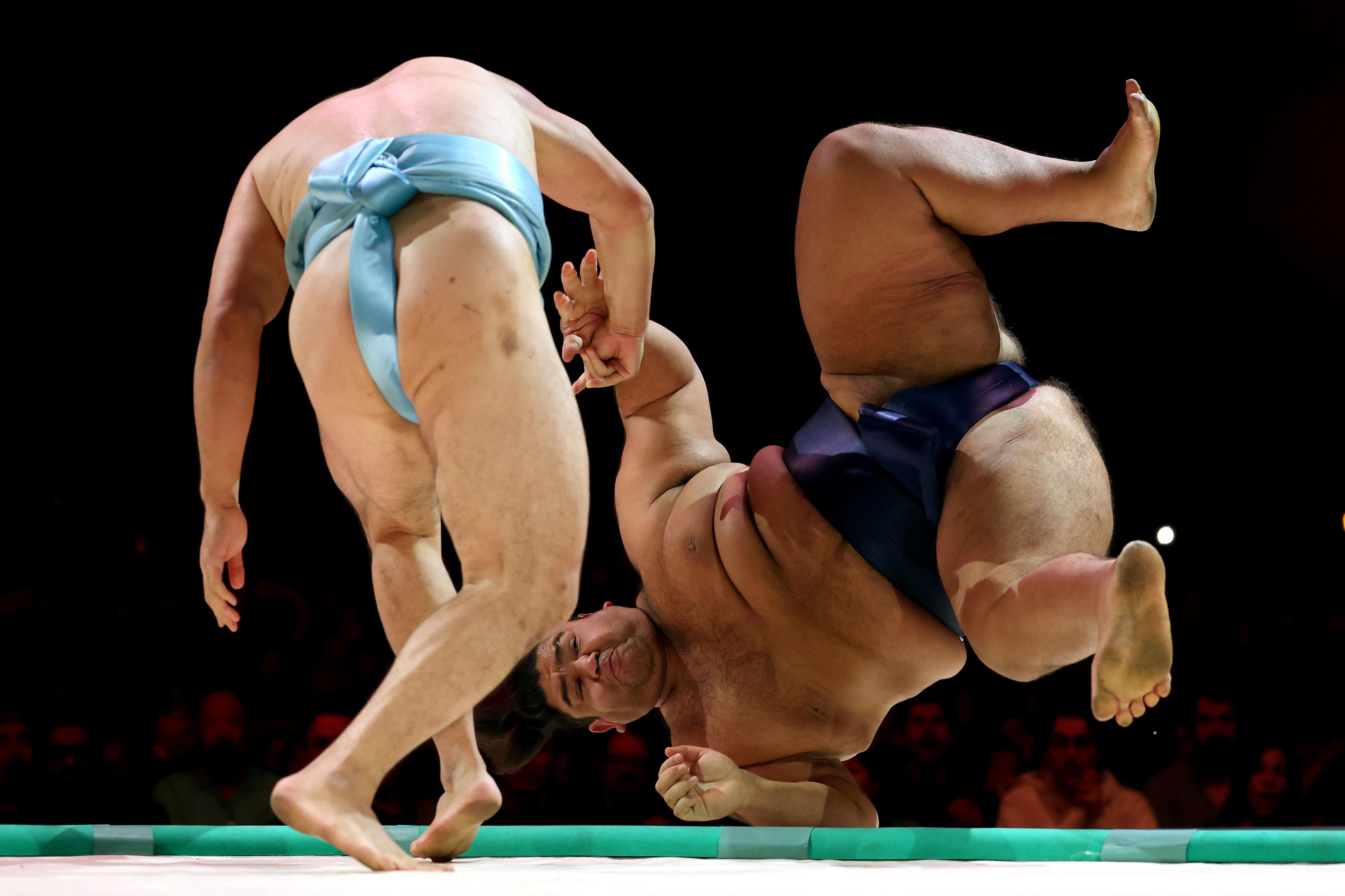 Two sumo wrestlers in a ring, one wearing a blue mawashi and standing, while the other in a dark mawashi is mid-air after being thrown, with dramatic lighting highlighting the action.