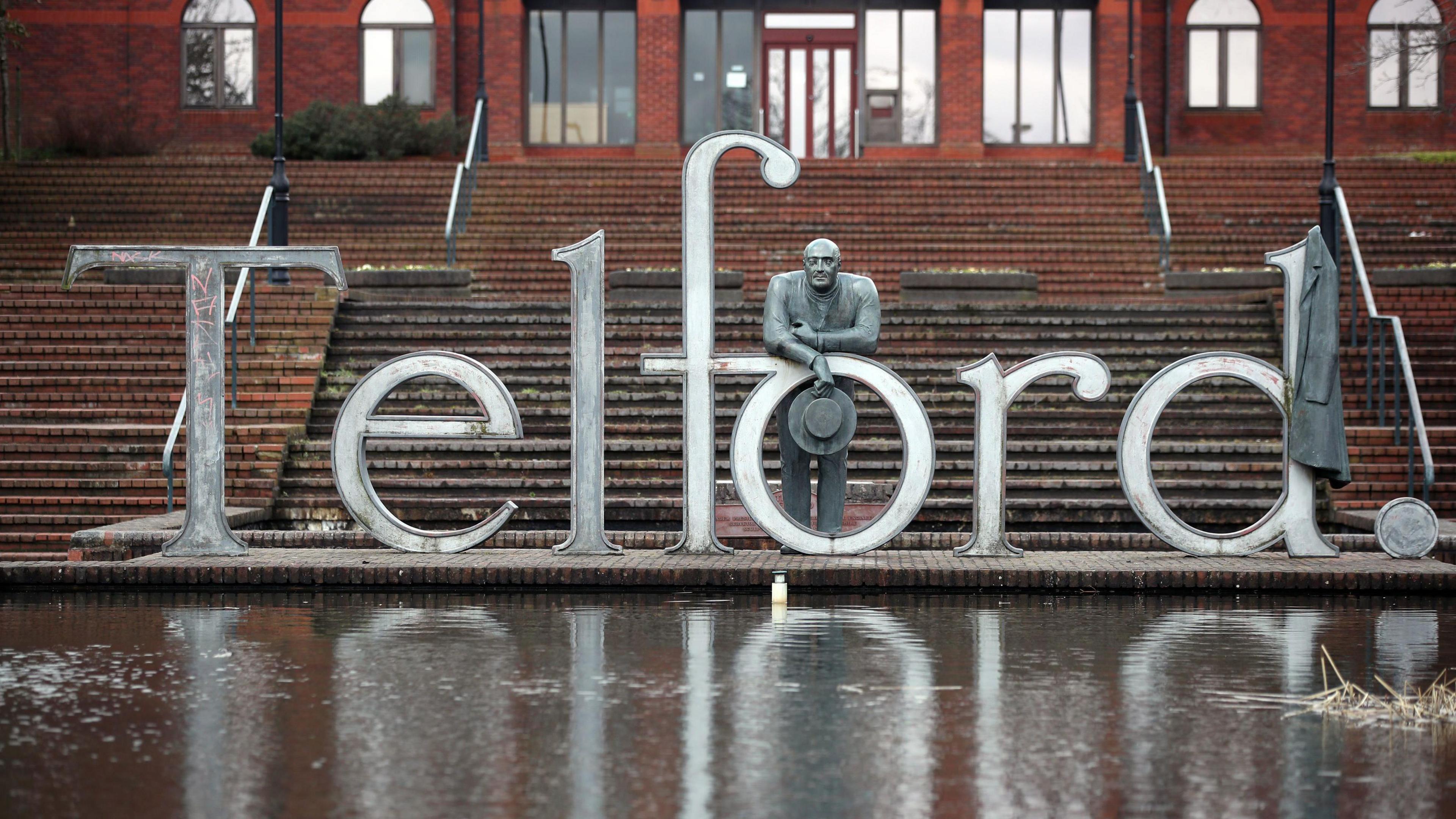 A large metal sign that reads "Telford" with a metal statue of a man holding a hat leaning over the "o", with brick steps behind it and a pond in front