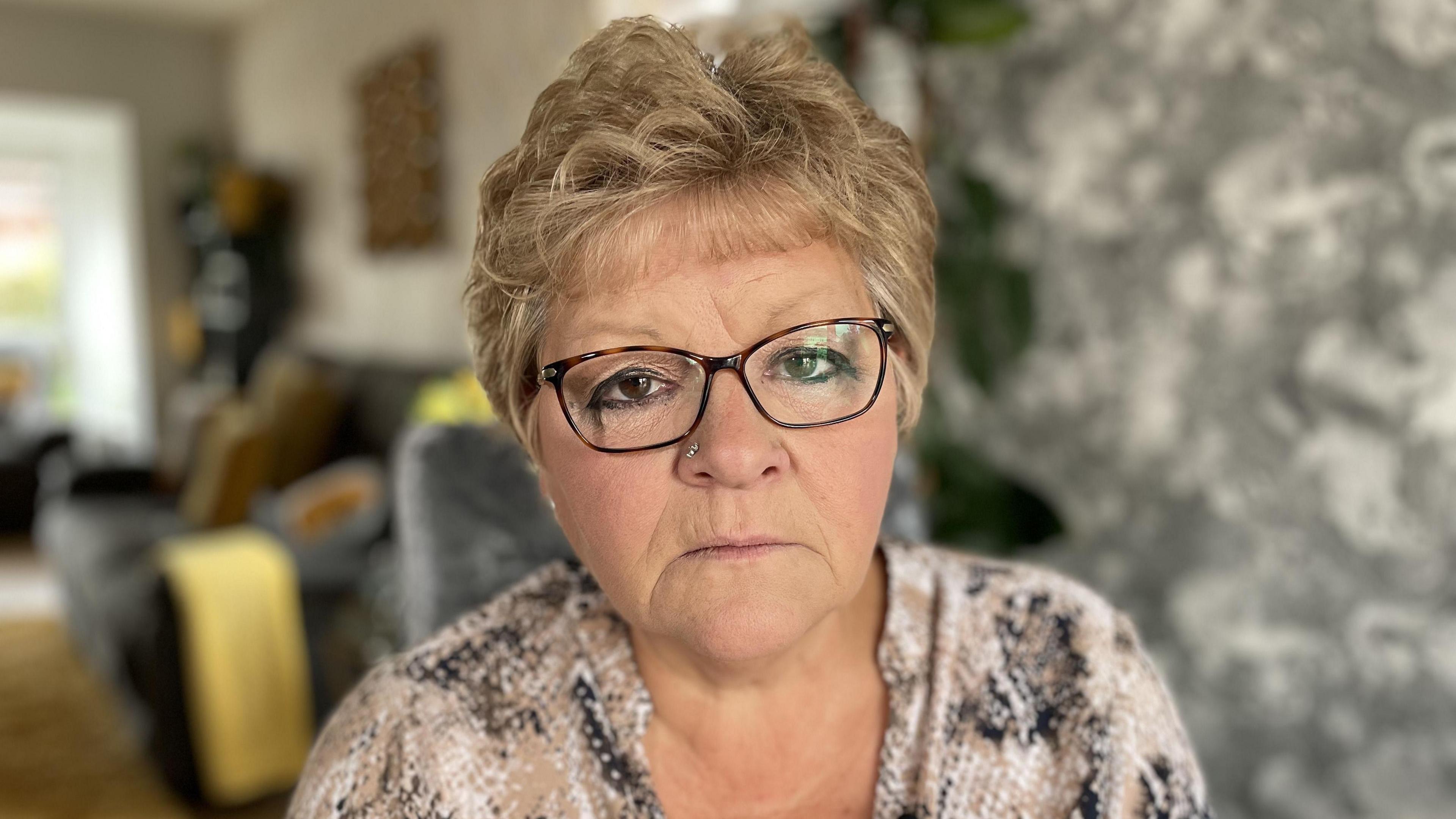 Tracy Canham stares at the camera. She has short blonde hair, dark eyes and thick-rimmed glasses. She is wearing a blouse covered in a snake skin print.