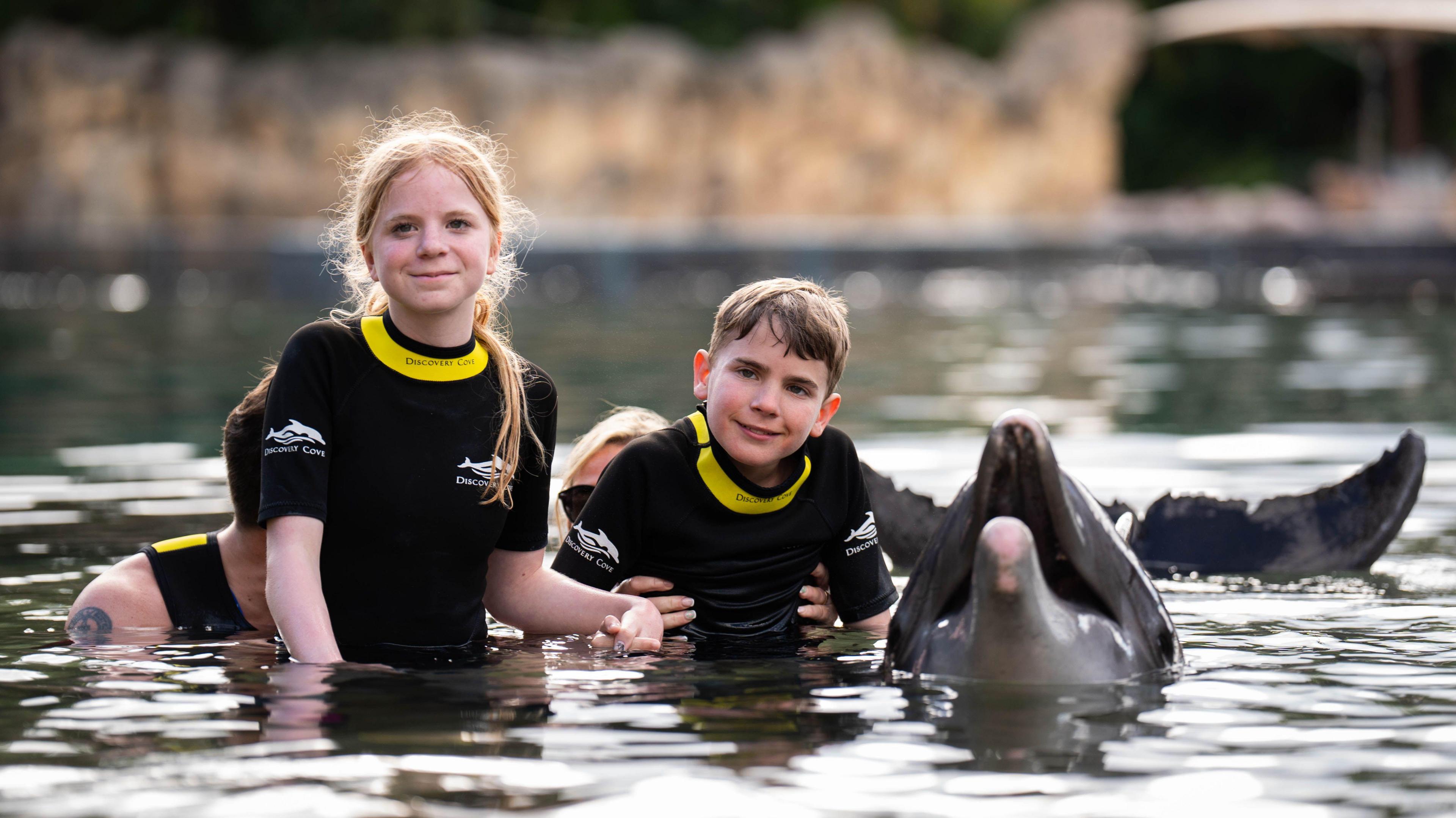 Lottie and Tommy are in black and yellow wetsuits in a dolphin pool. They are holding hands, and next to them is a dolphin which has its mouth open