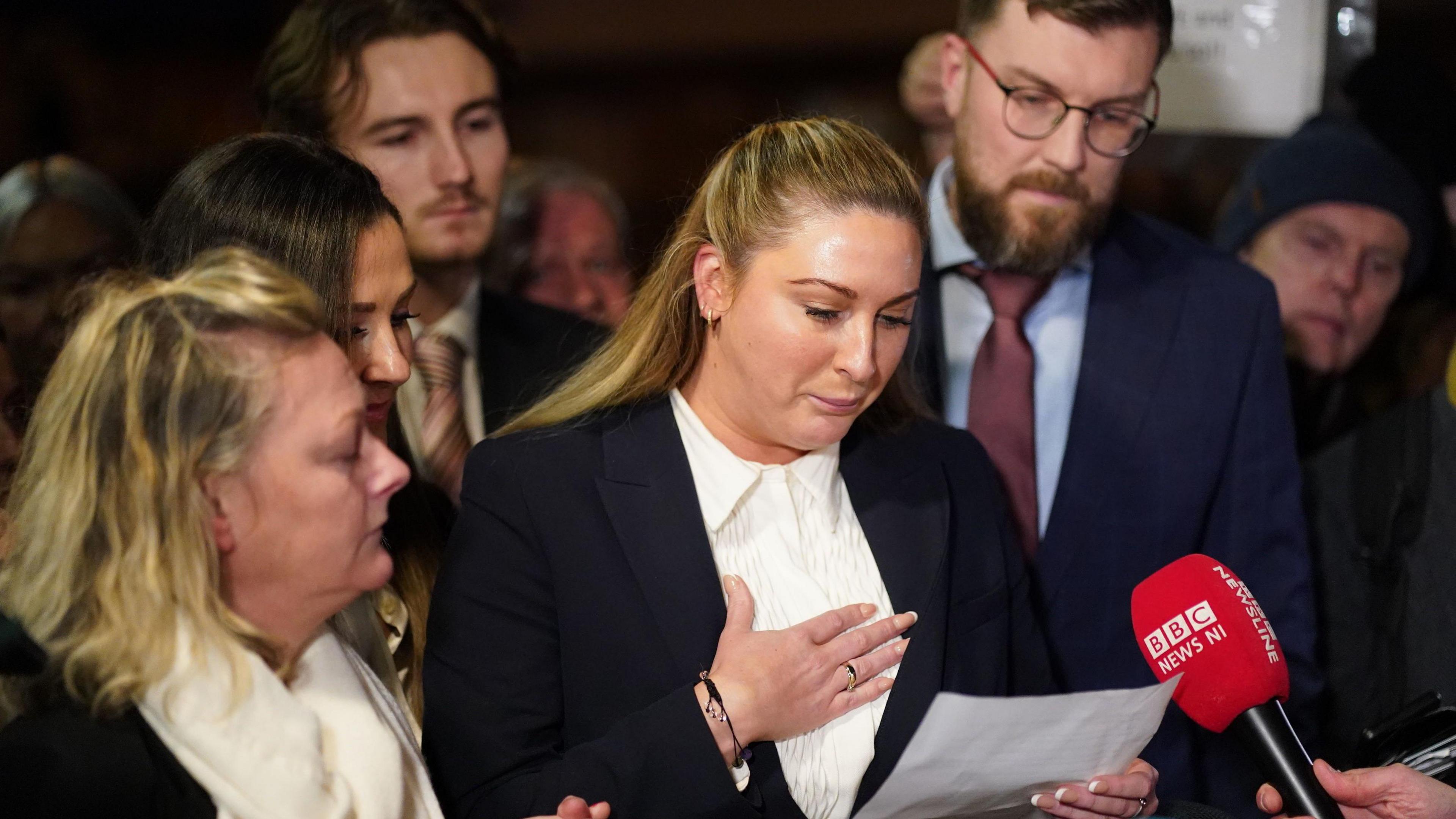 Nikita Hand, surrounded by two men and two women. She has blonde hair and is wearing a white top and black blazer. She has her hand on her chest and is looking at a piece of paper in her other hand. There is a red BBC News microphone held up in front of her. 