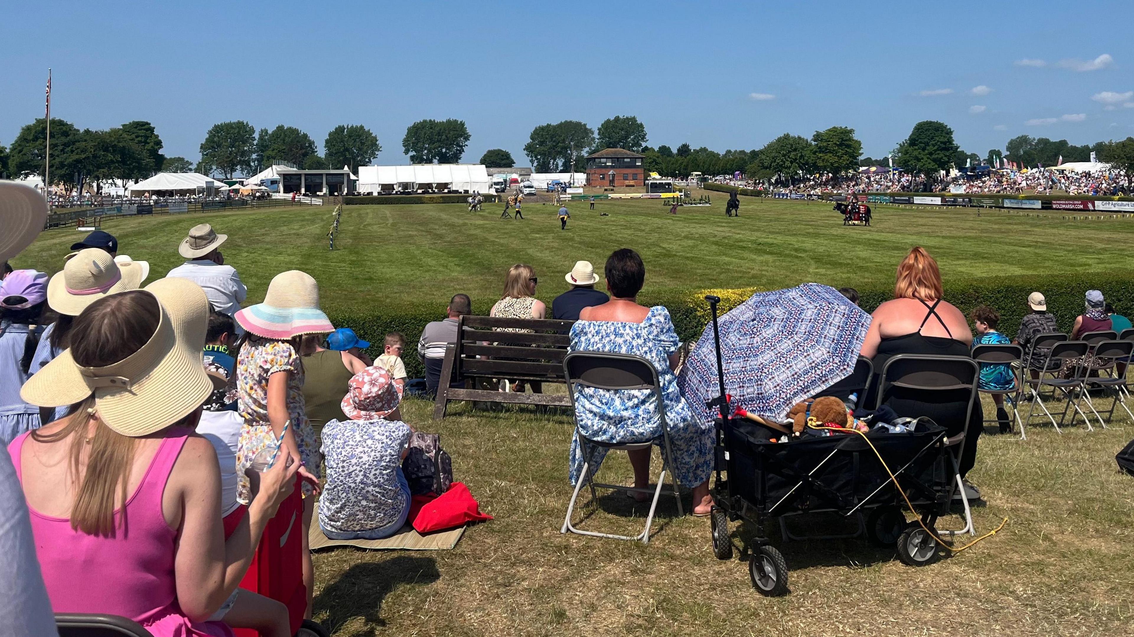 A crowd of people - many wearing hats - seated around an event ring. There are horses and jumps in the background.