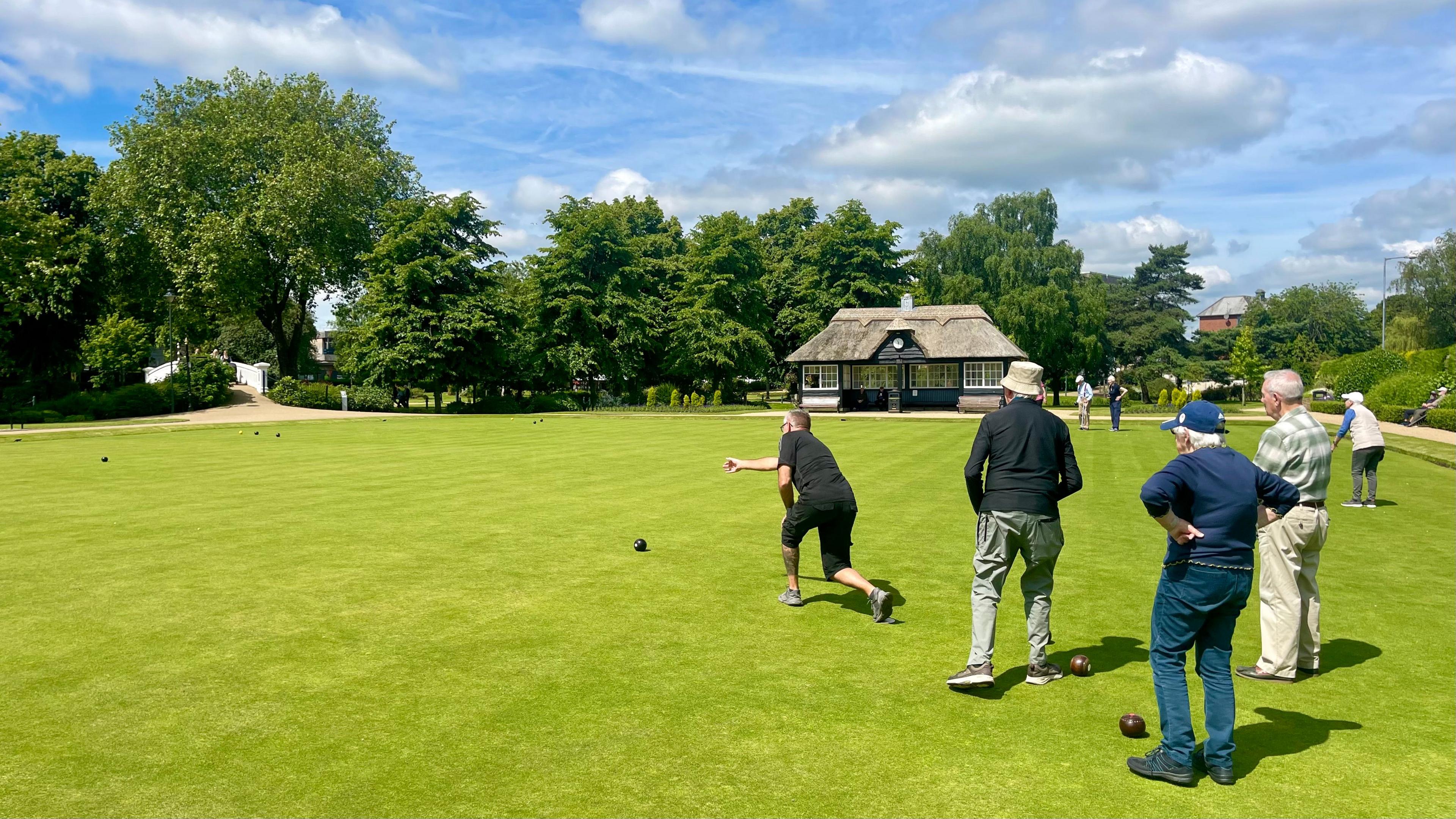 Five men are playing bowls on a sunny outdoor green. One man in a black T-shirt and shorts is releasing a ball. Three others watch his throw, two of whom are wearing sun hats. A thatched seating area with a clock face stands at the top of the green with large thick trees in the background. A sandy pathway surrounds the bowling green.