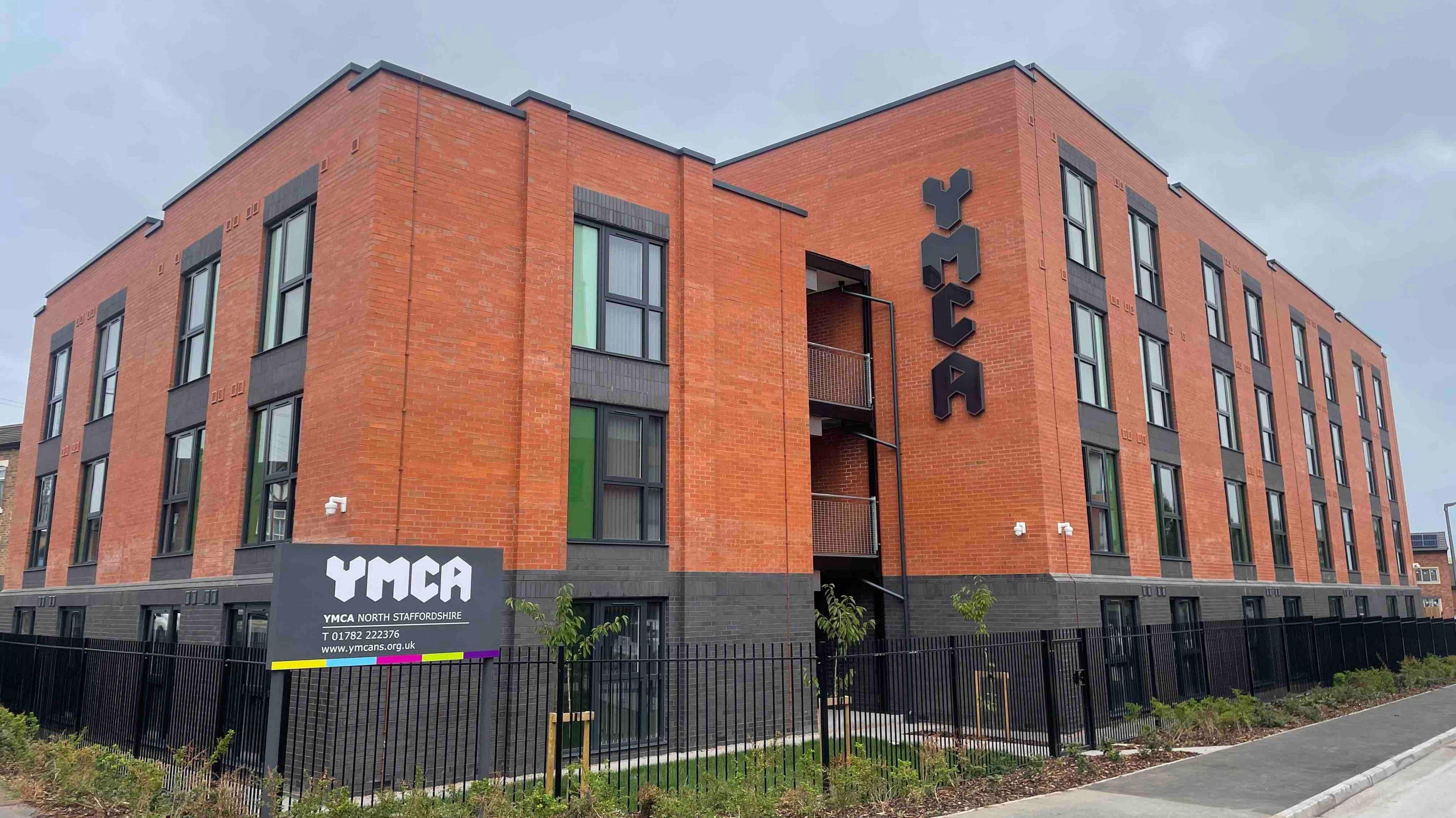 A brick building, behind black metal railings. There are two-storey and three-storey sections of the building. One wall has black lettering on it which reads YMCA.
