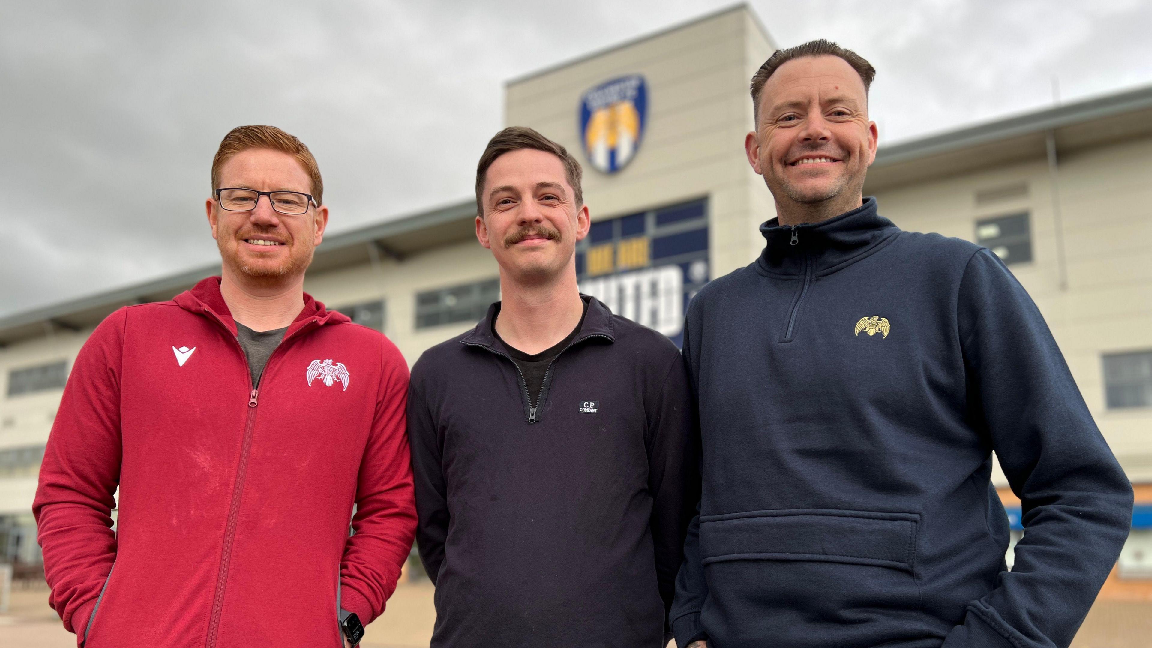 Colchester United fans (left to right) David Clayton, Aaron Jay and Si Collinson. All three are standing in front of the Colchester United stadium. David wears a red zipped hoodie with white logos on both chest panels. He has ginger hair, a stubbly beard and silver-rimmed glasses. Aaron wears a black quarter-zip top. He has short brown hair and has a moustache. Si wears a navy quarter-zip top. He has short dark hair. All three are smiling at the camera.