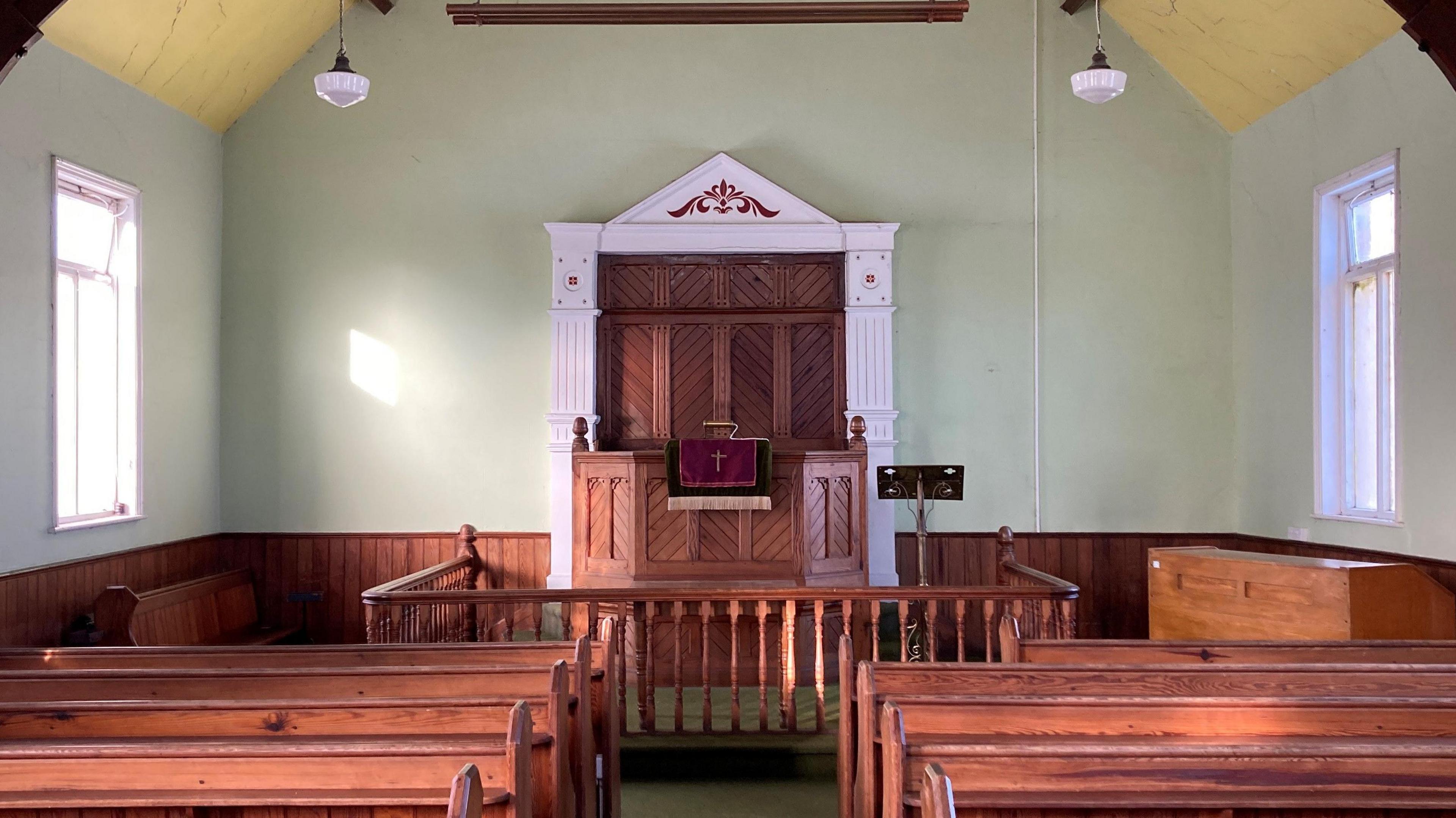 View of the inside of the chapel. Pews on either side with an ornate pulpit in the centre on the far wall. There's a piano or organ on the far right side.   