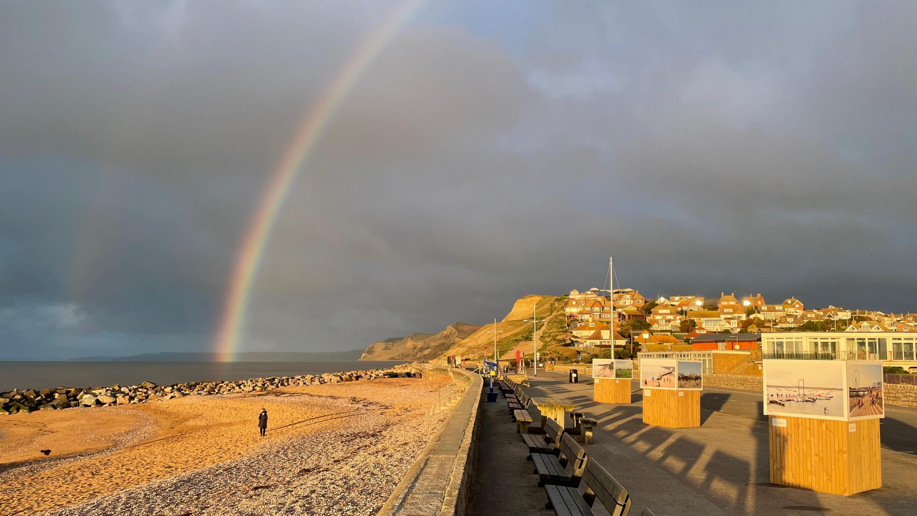 Two rainbows can be seen, one much brighter than the other, ending in the sea. There is a lone walker on the beach which is flanked by a row of wooden benches. Cliffs can be seen in the background alongside several houses.