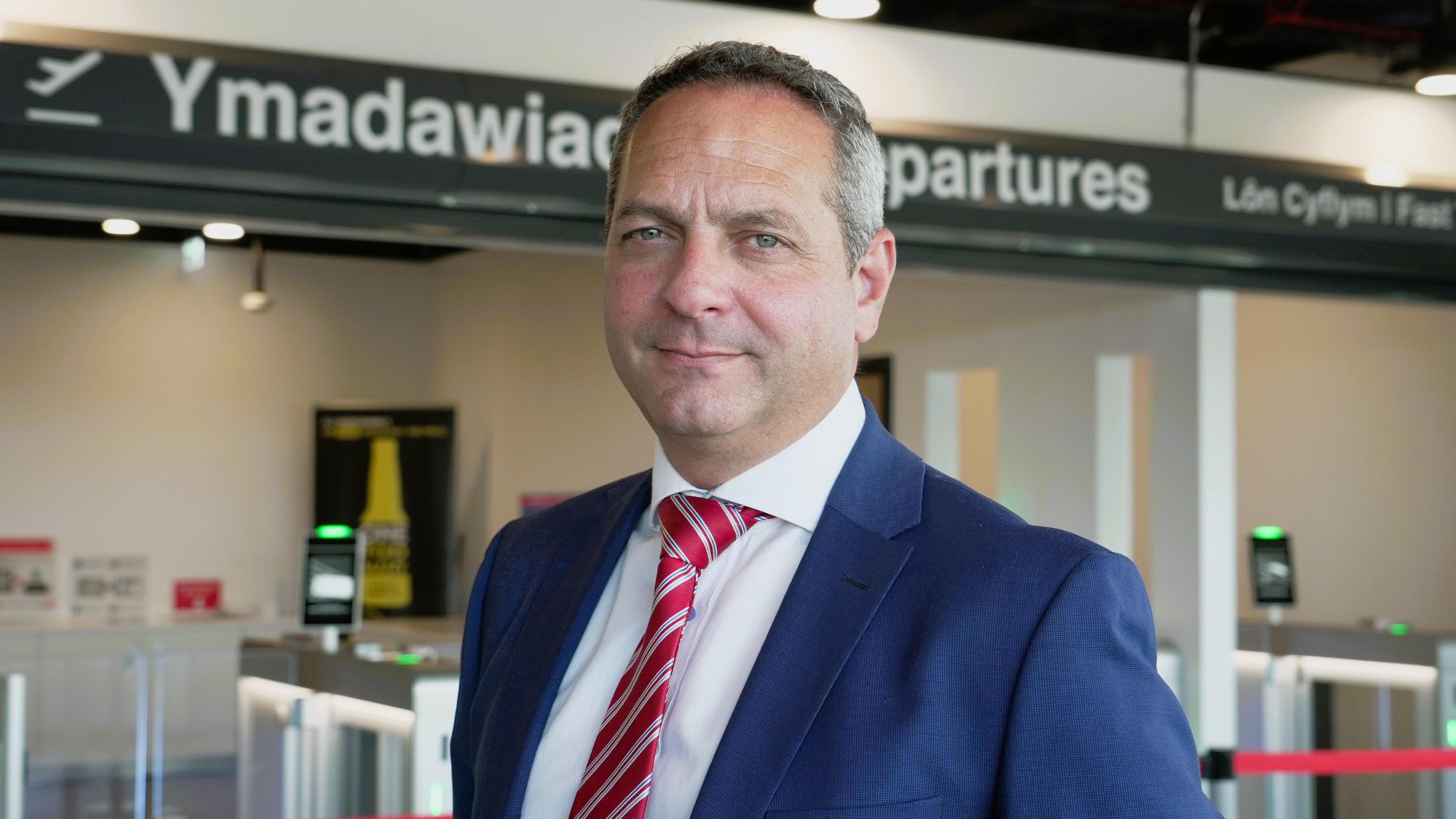 A headshot of Spencer Birns, chief executive of Cardiff Airport, wearing a blue suit and red tie. He is standing in front of the security barriers that take passengers to the departure gates.