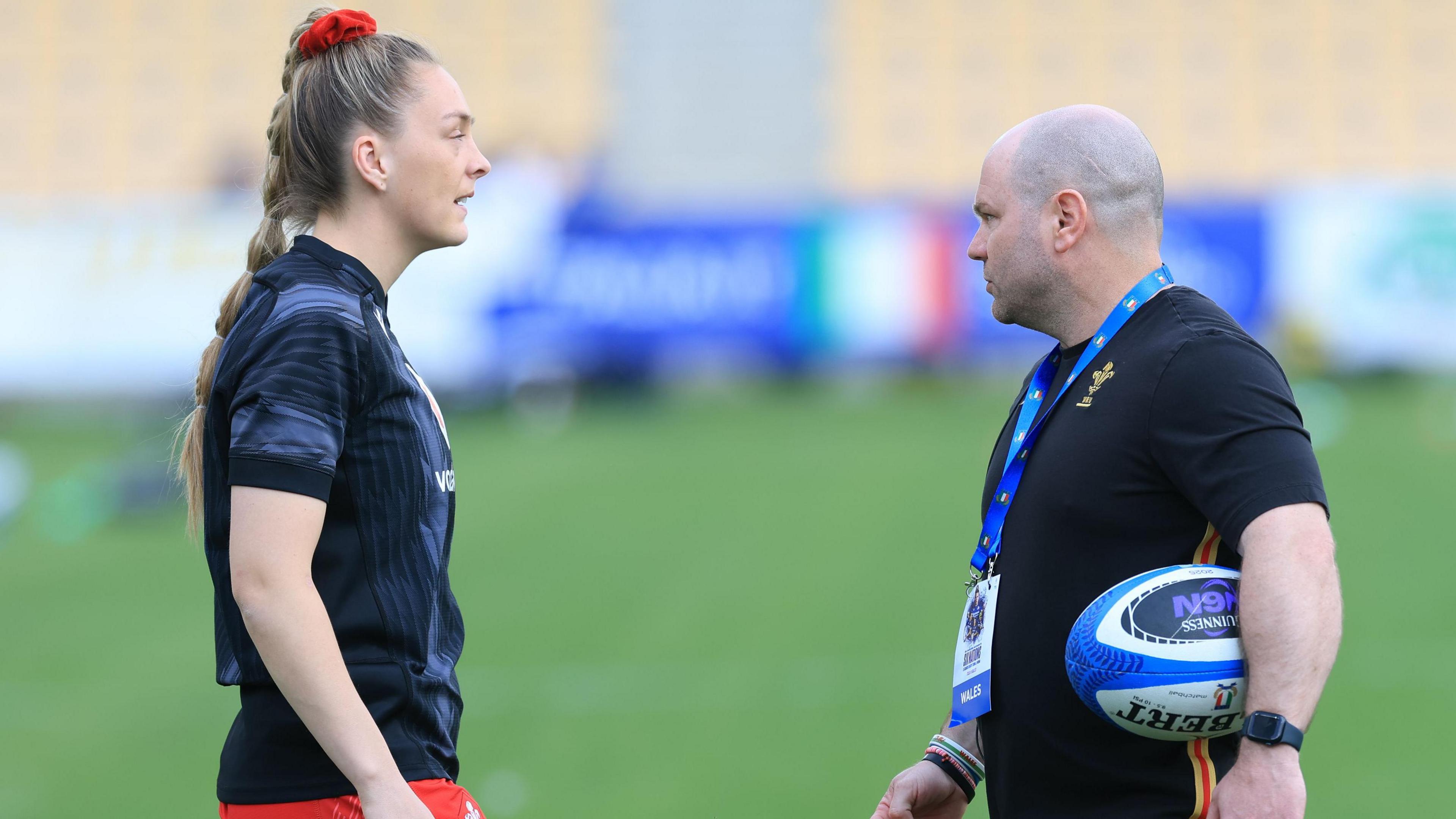 Hannah Jones in a training top talks to Sean Lynn who has a rugby ball under his arm on a pitch before a game.