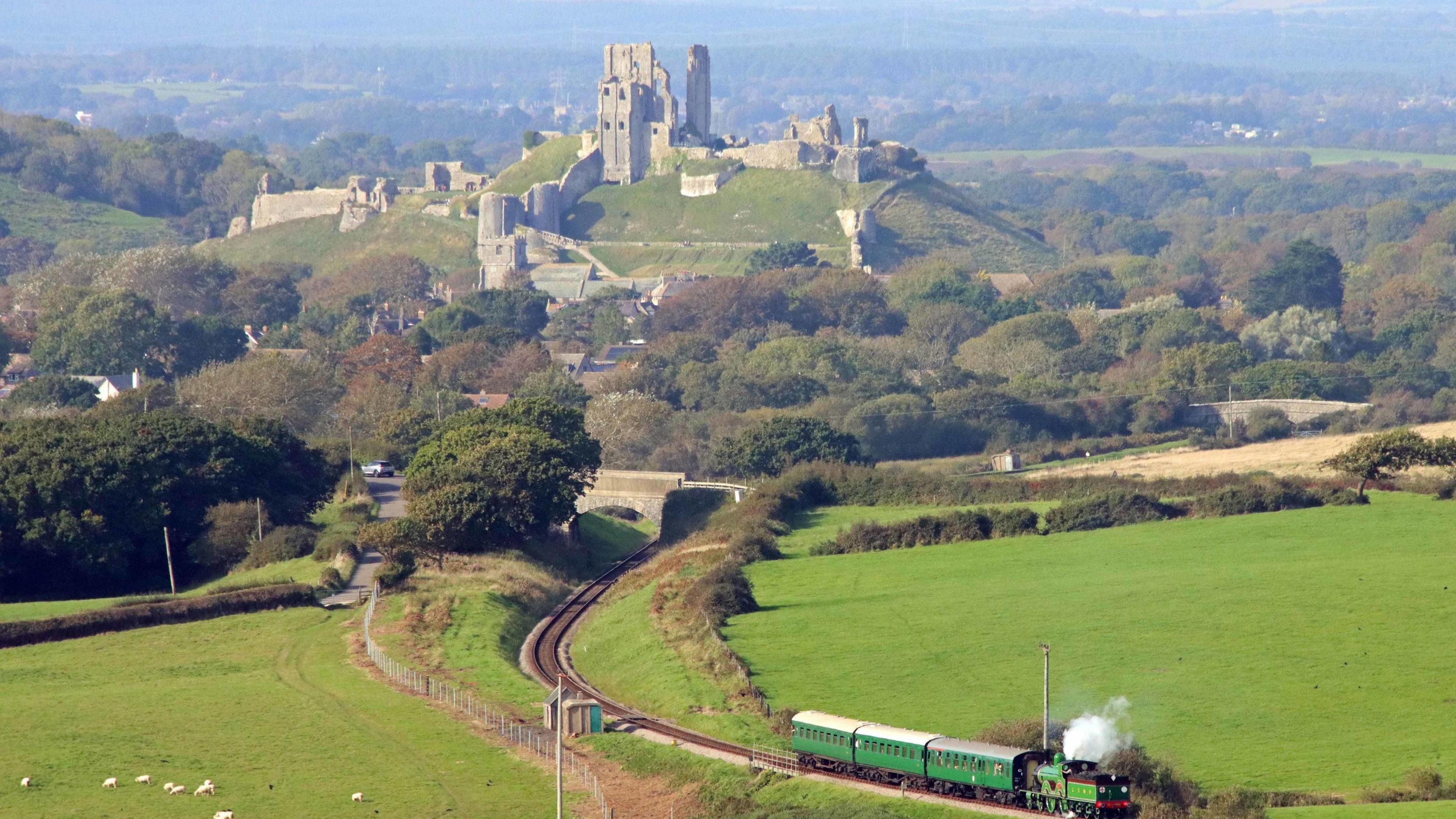 Corfe Castle photographed from far away on a sunny day. A Victorian steam train can be seen passing through the foreground