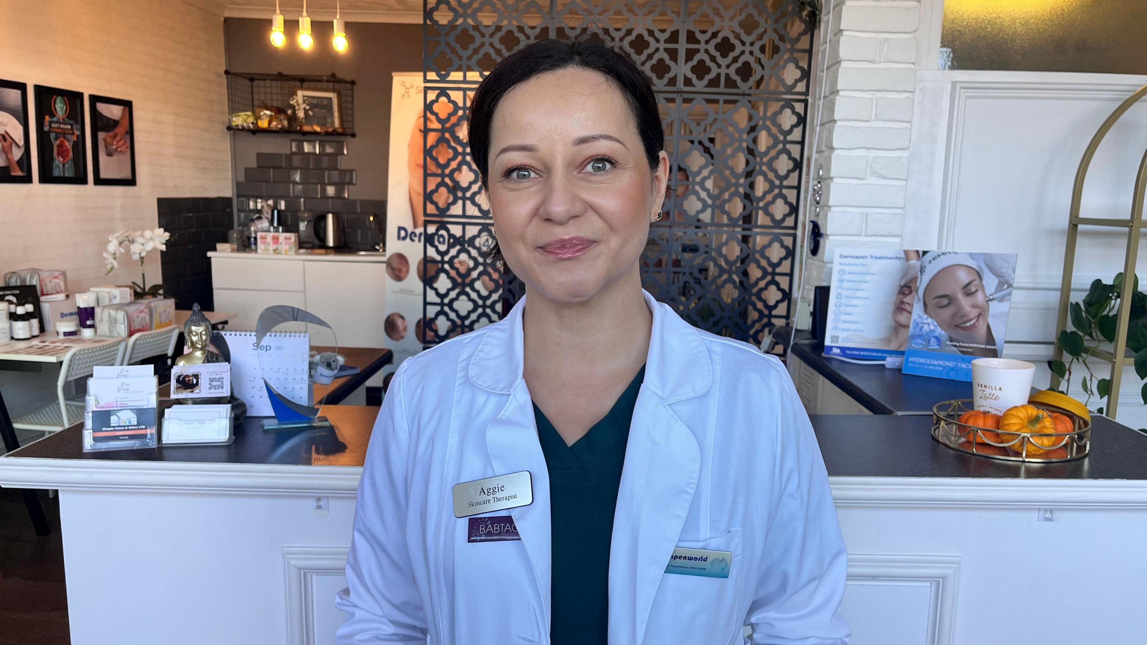 Aggie Singh stands in the reception area at her beauty salon. She is wearing a white jacket and black top and has dark hair, which is tied back. Behind her is a white counter with a black top, covered in paraphernalia for the salon. A decorative screen, artwork and soft lighting can be seen in the background.