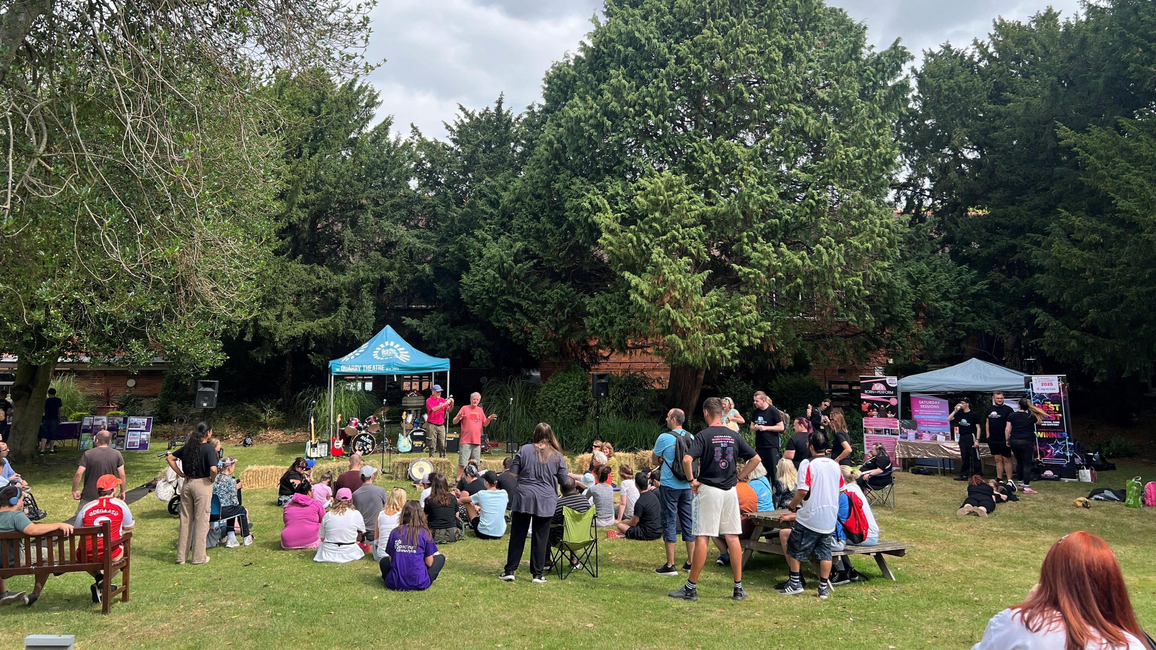 A large group of people outside in a garden watching a performance, with gazebos in the garden. Some are sitting down, others are standing up. There is grass and trees in the distance. 