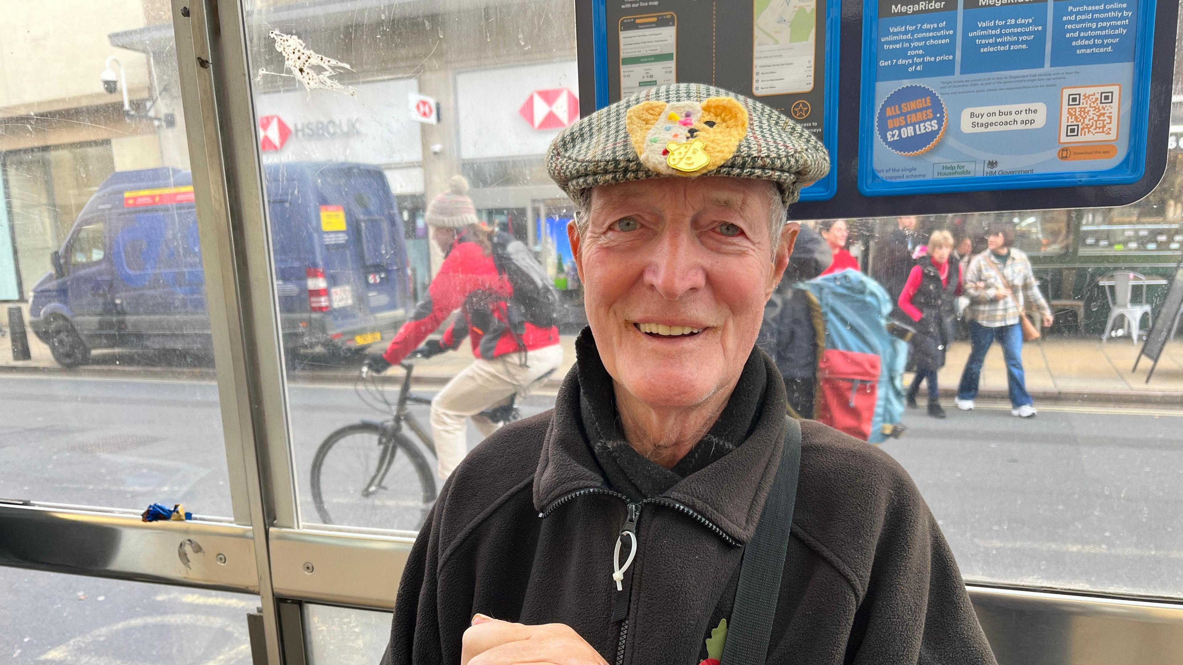 Trevor Minchley is sitting under a bus shelter, smiling at the camera. He is wearing a flat cap with a Children in Need Pudsey badge on it and a black fleece.