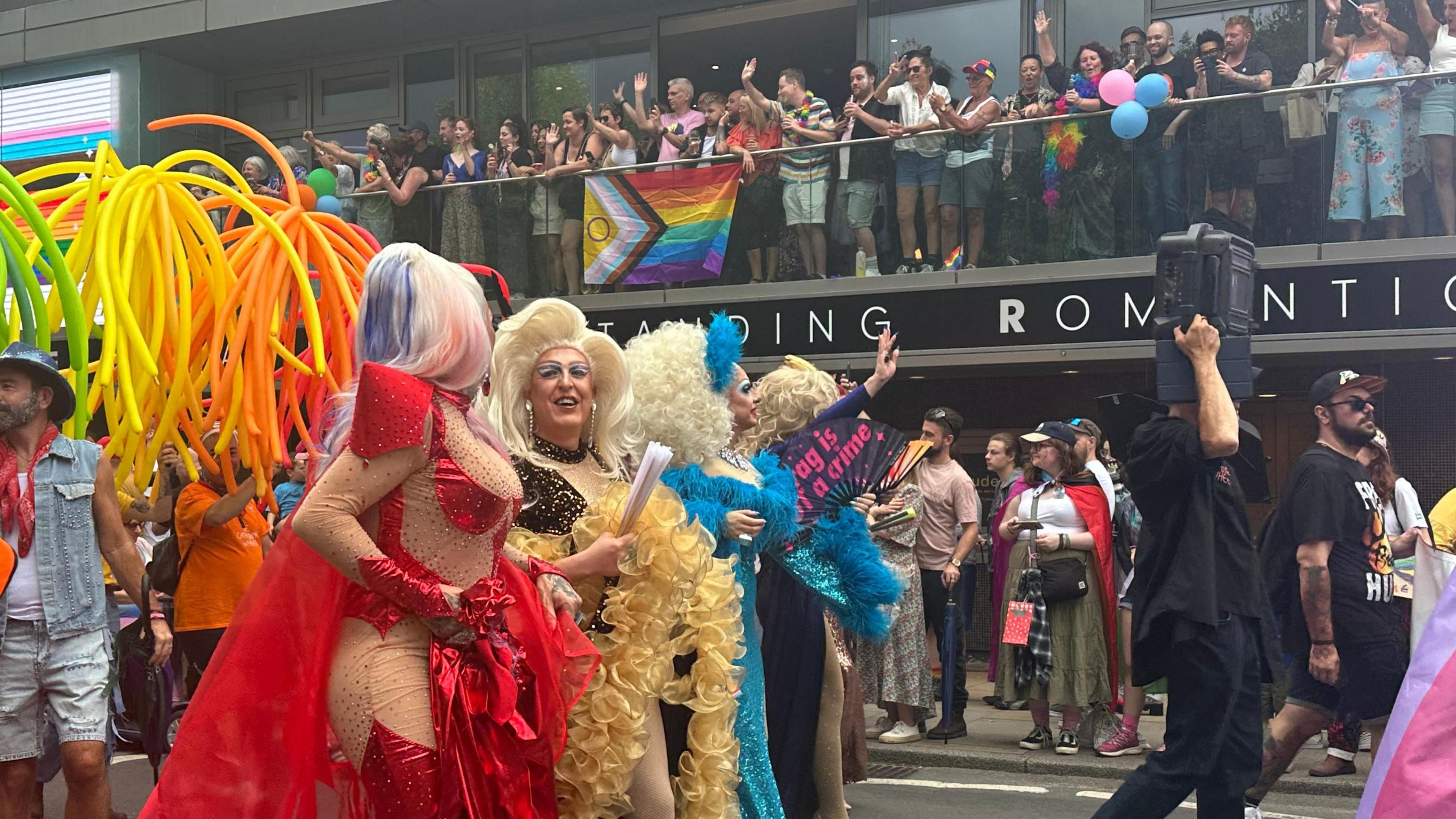 Drag queens are standing on the left walking across the image. They are all wearing different coloured outfits, and people are walking beside them in the same direction. A building is behind them with people standing on a balcony. 