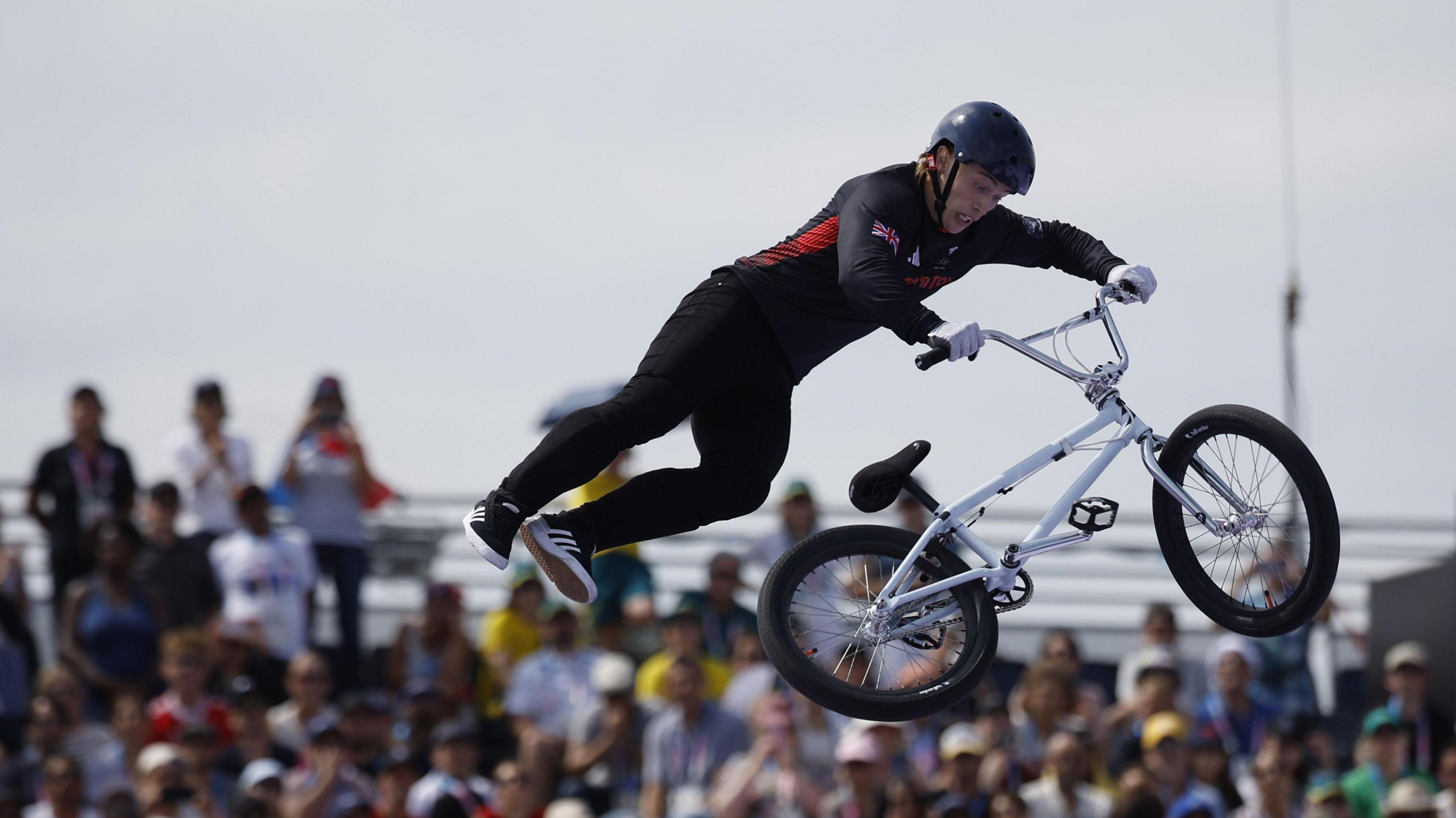 Kieran Reilly in the air performing a trick at the Paris Olympic Games. His legs are in the air as he spins the white bike. The crowd can be seen in the background.