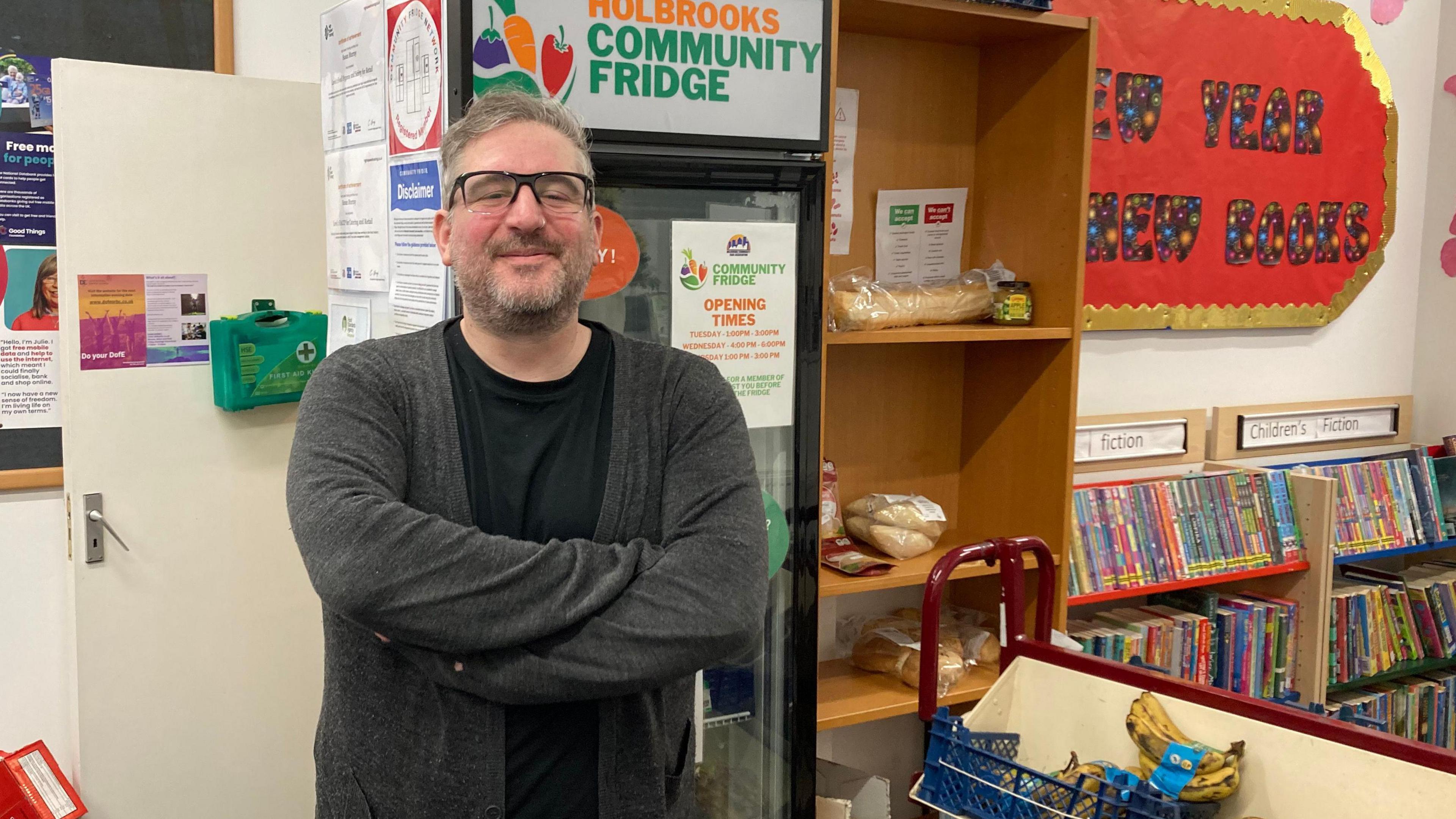 Mark Graham standing in front of a fridge in a room which has shelves of books and shelves containing fruit and bread. He is wearing a black top and dark grey cardigan and has his arms folded in front of him. 