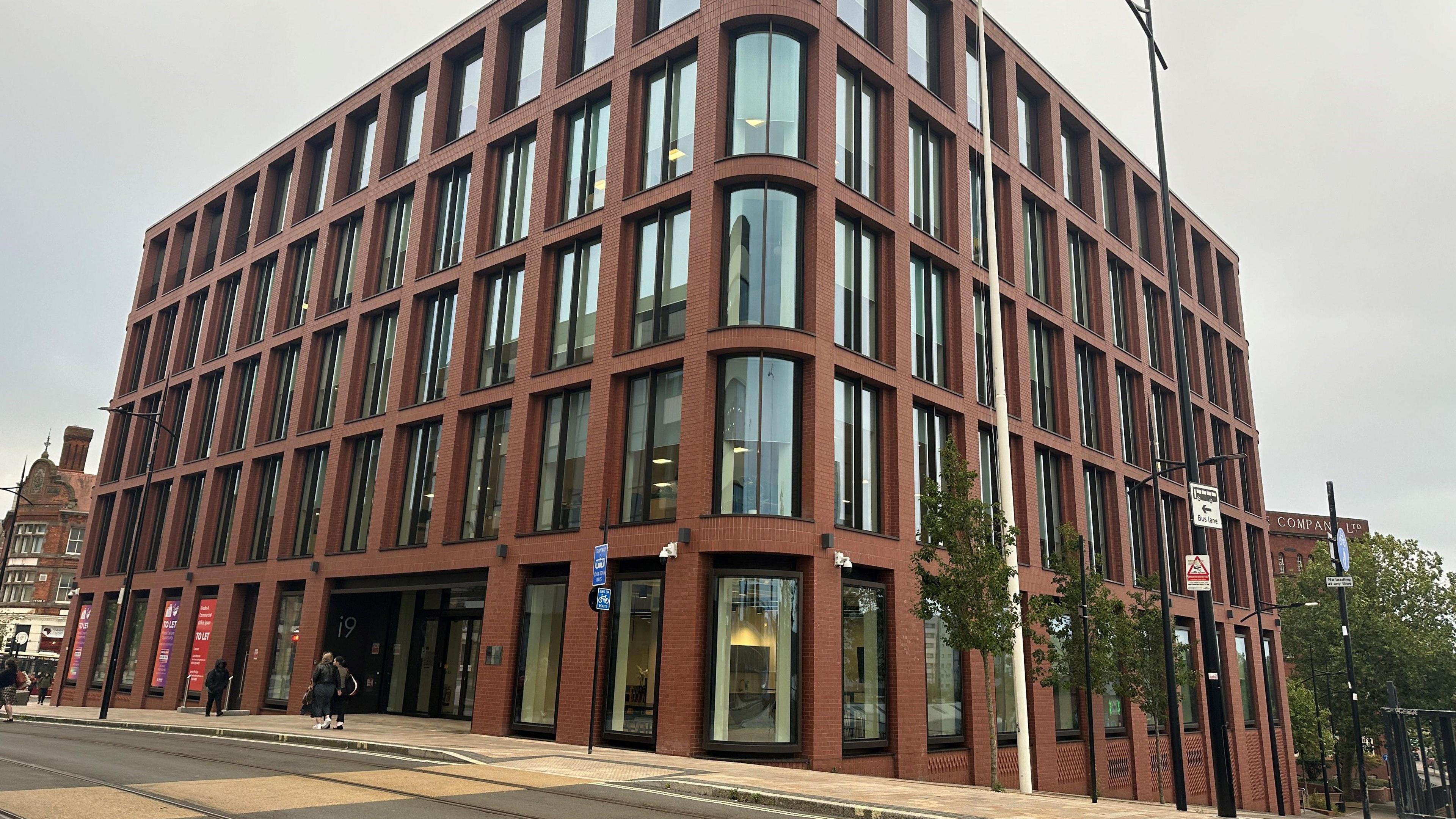 A modern five-story, brick building with extensive glazing, including curved windows on the corner facing the camera. 