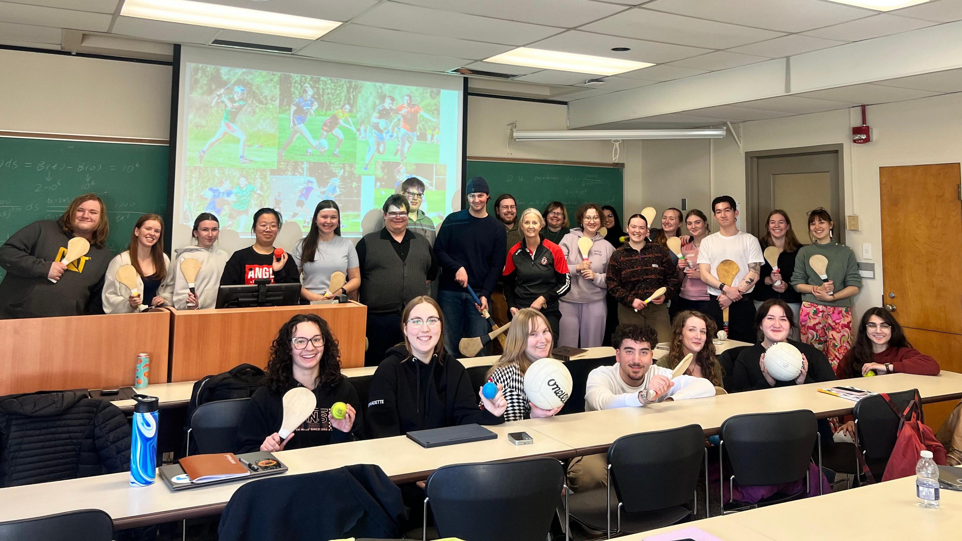 26 young people stand in a classroom smiling at the camera. They are learning about Irish sports and holding various pieces of equipment.