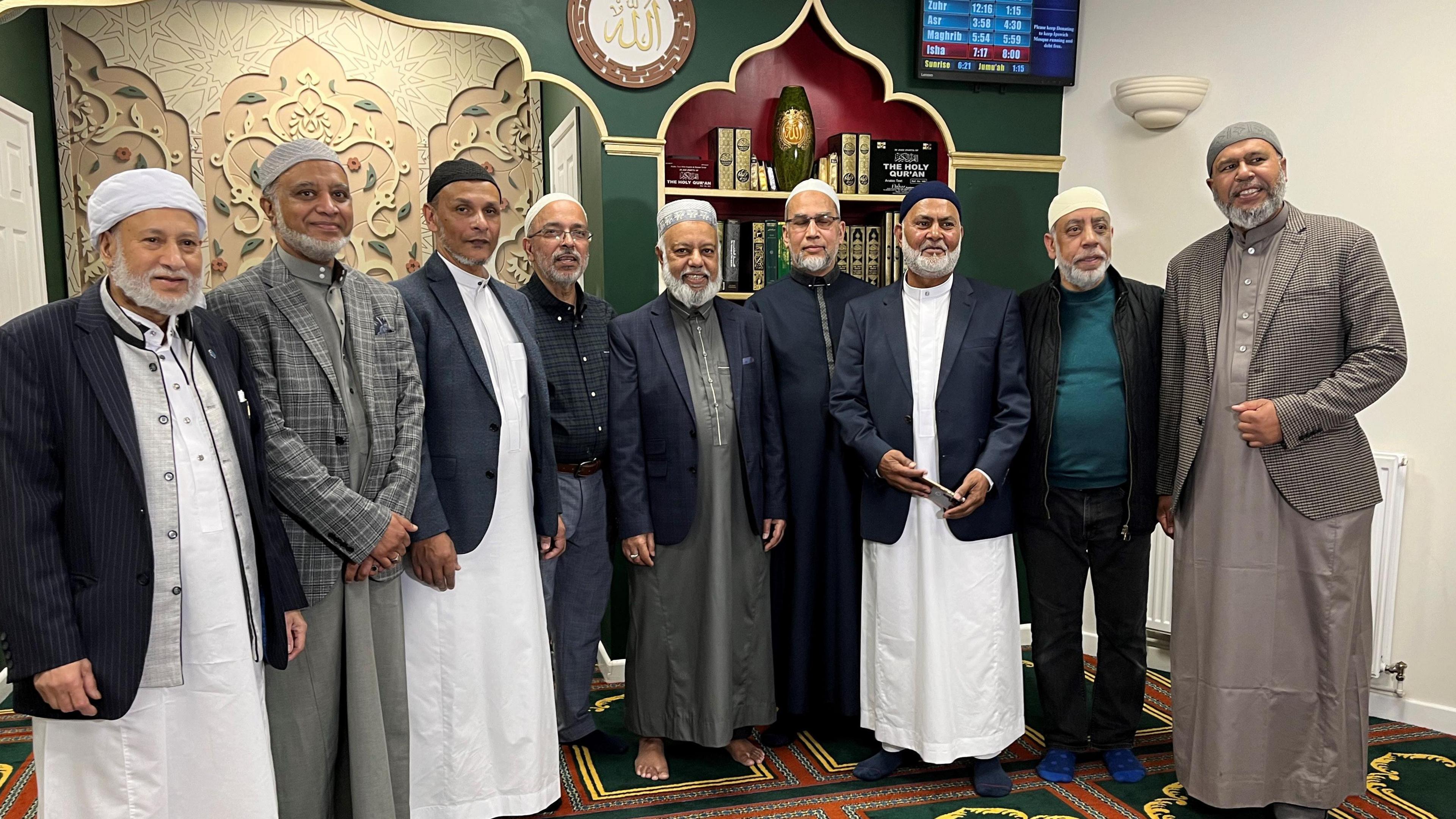 Nine men standing in a line at the front of a mosque. They are wearing traditional Muslim robes (called Thobes)