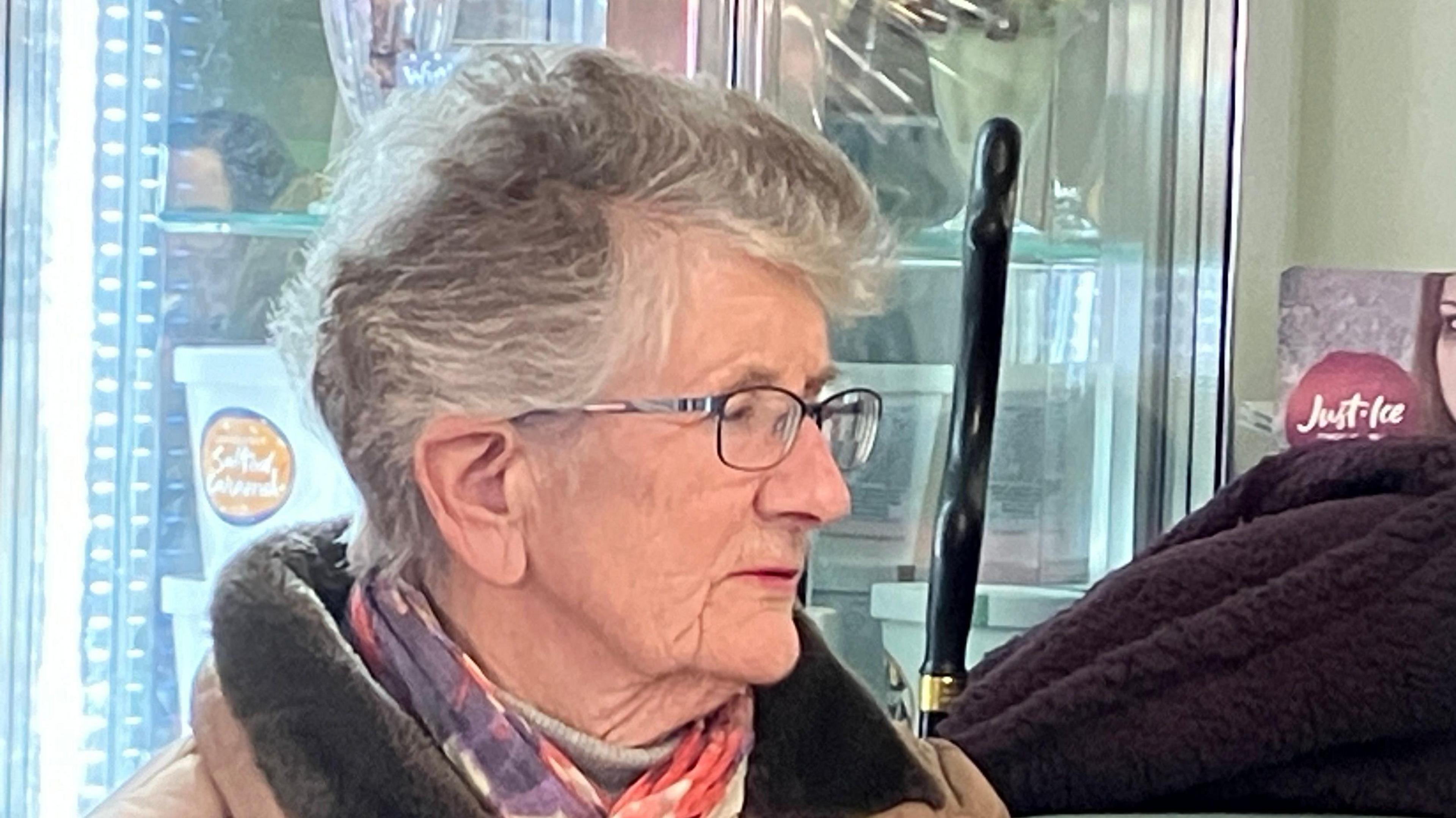 An elderly lady with grey hair and wearing glasses is interviewed in a cafe. She appears side-on to the camera.