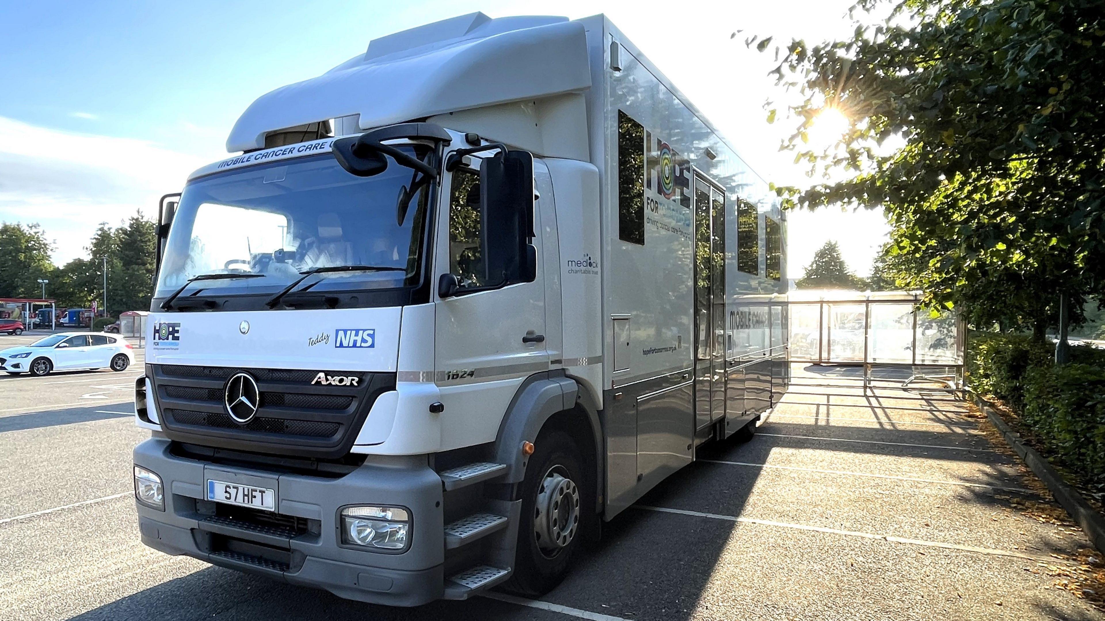 A white truck parked at a supermarket car park. On the front of the truck is the NHS logo and on the side is a large door and windows
