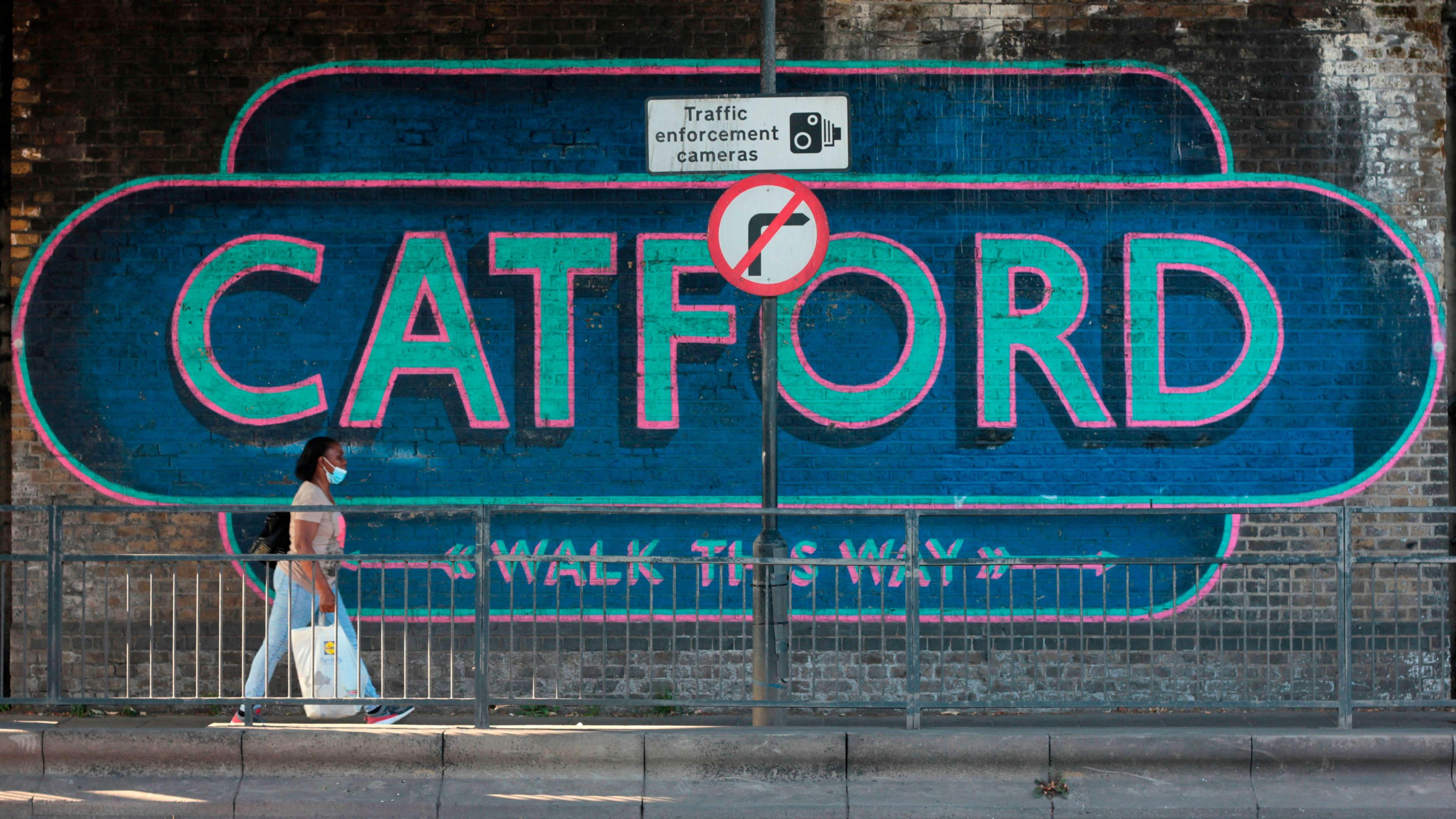 A blue mural with the word 'Catford' behind a woman walking past and a traffic enforcement cameras and no right turn sign
