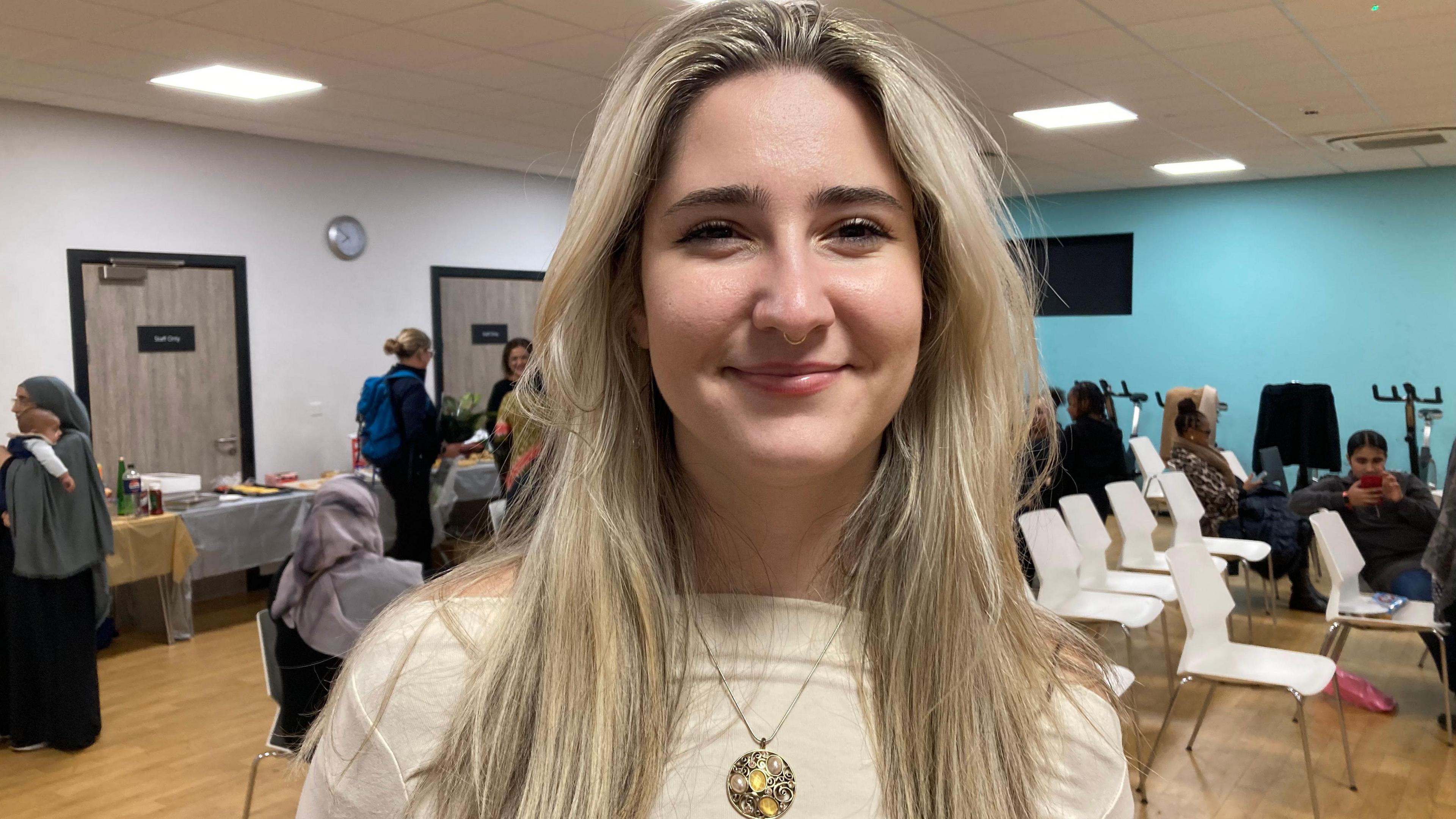 Rafaela Marinkovic stands in a room with other group members around her. She is wearing a cream top and necklace and has long, blonde hair. Other people are standing and sitting in the background and talking.
