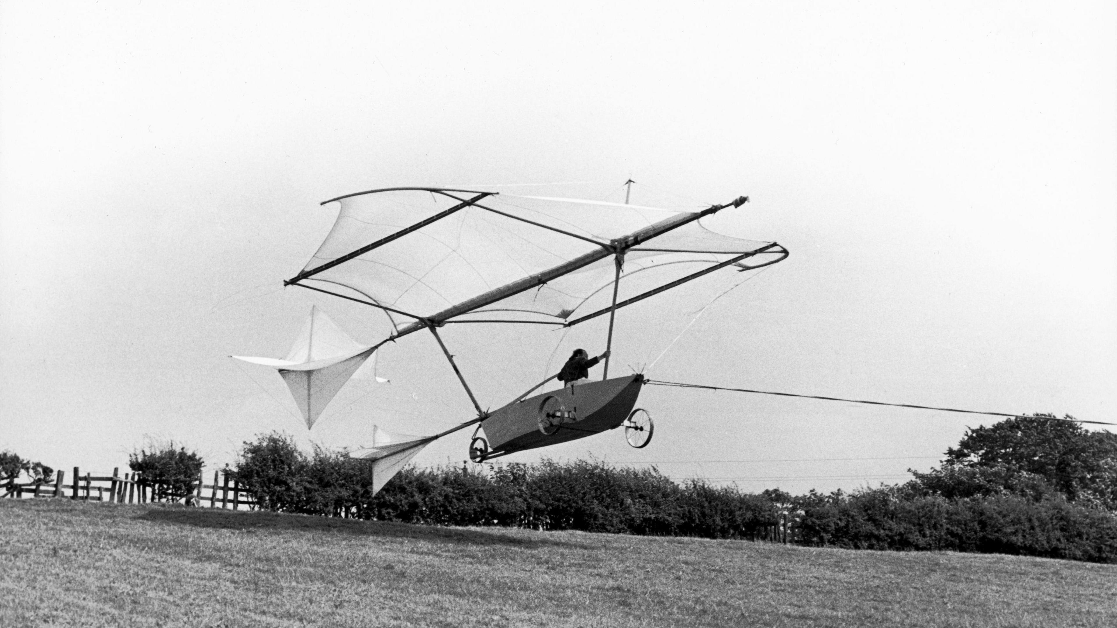 Black and white photo depiction of Sir George Cayley's flying machine in the air over a grass field.  The aeroplane has a canvas wing above a canoe-like shape with three wheels.