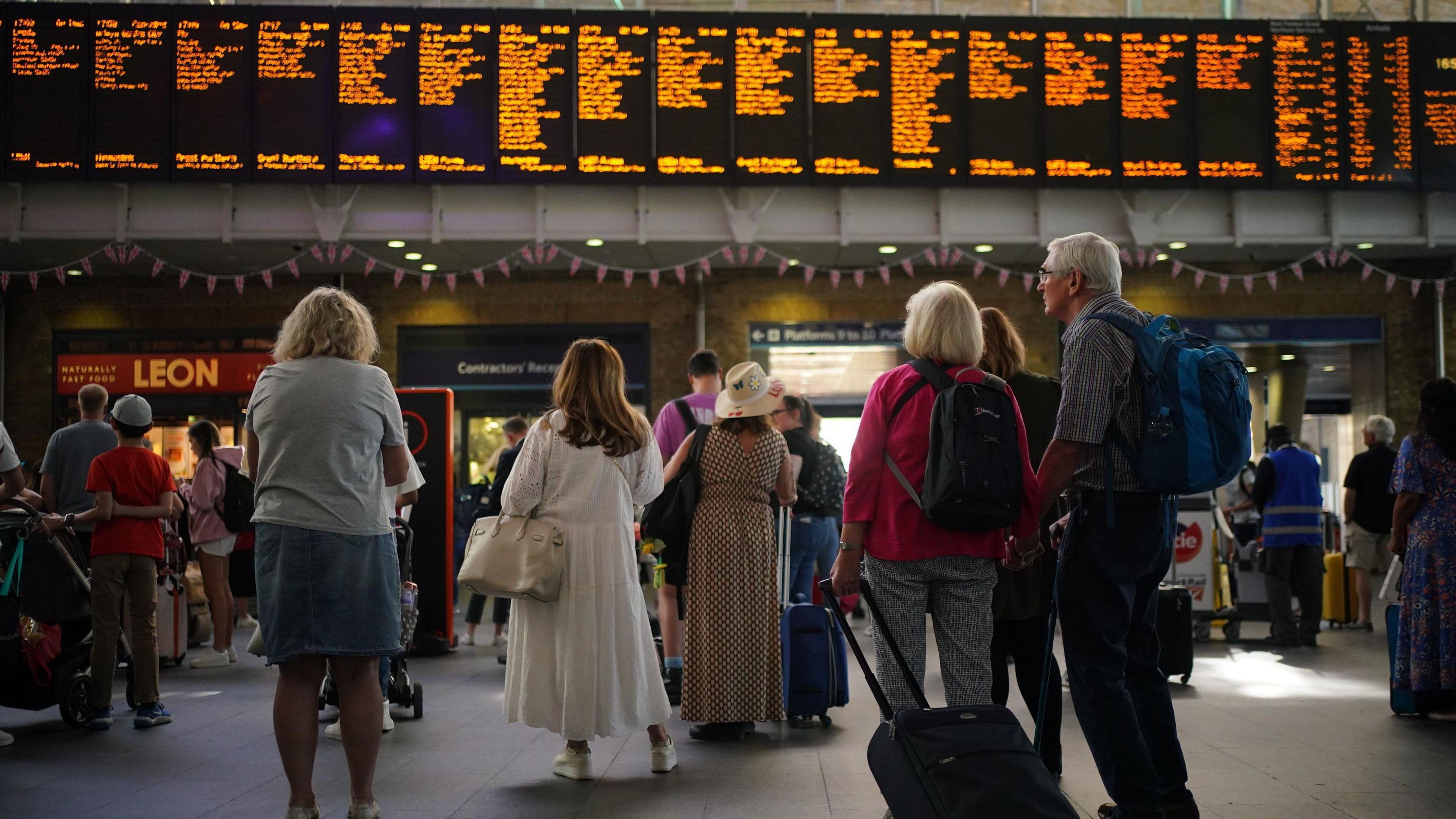 A group of people stand with their backs to the camera looking up at an electronic departure boards in a busy London train station. An older couple to the left have backpacks and a suitcase. 