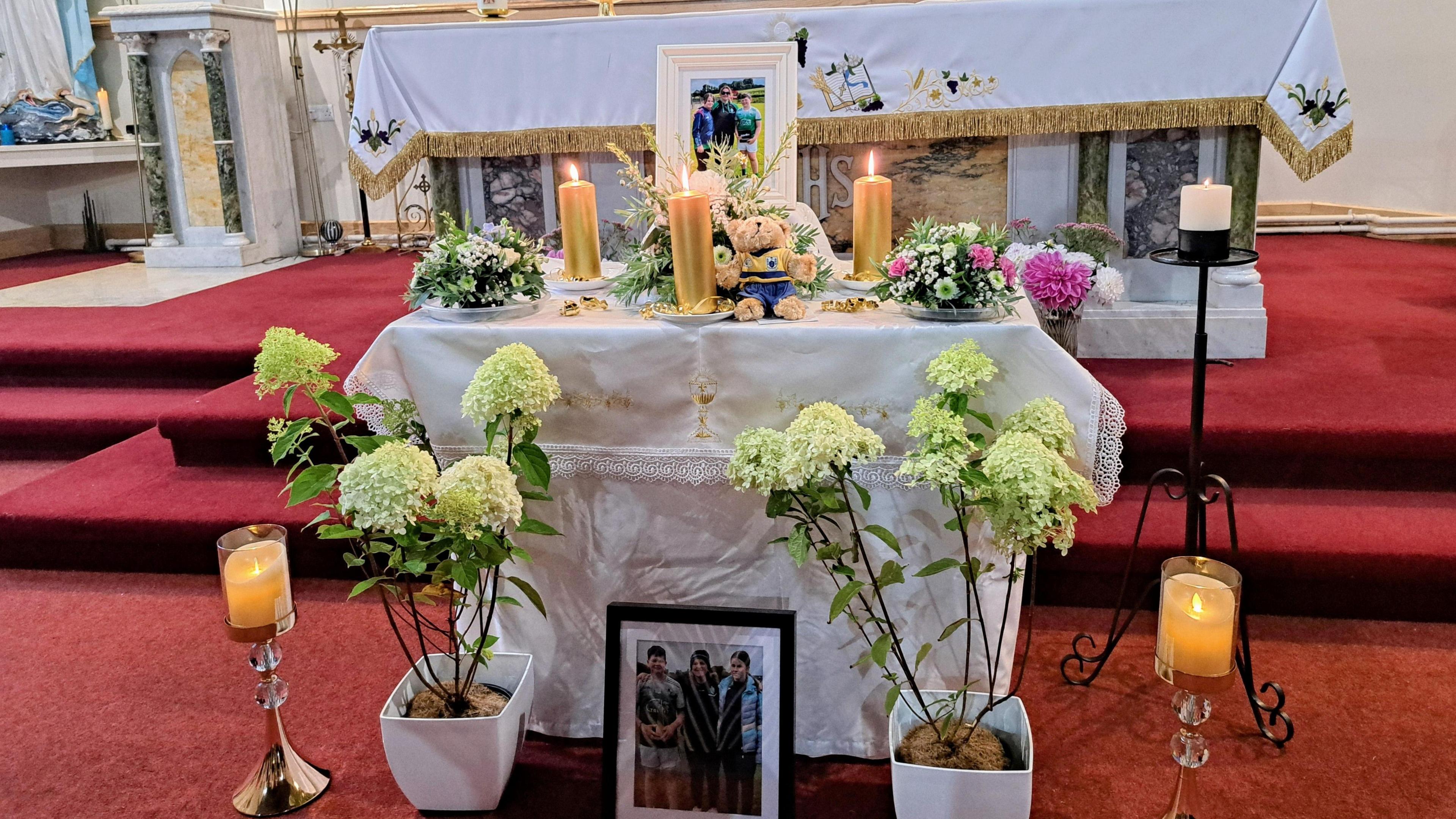 The interior of the Church of the Immaculate Conception in County Clare. Three candles are lit on an altar table which has been placed at the foot of red carpeted steps. There are photos of a woman and two teenagers on top of and beneath the altar. A teddy has also been placed on the table. Flowers surround the candles.