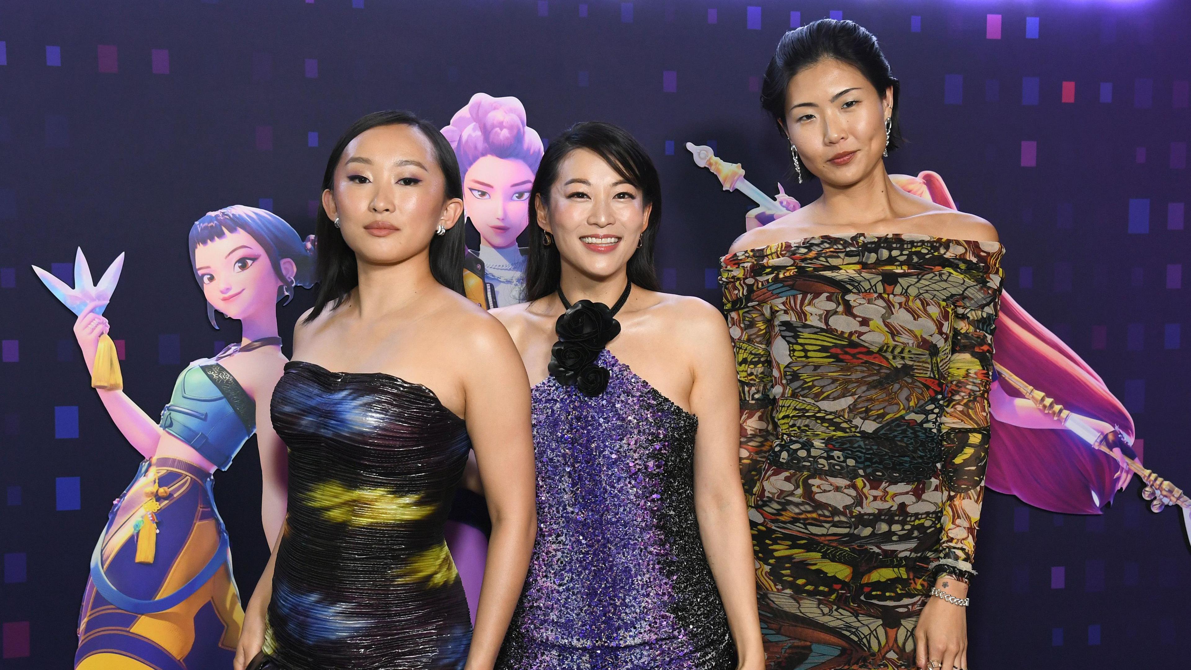 Picture of the stars of KPop Demon Hunters - Ji-Young Yoo, Arden Cho and May Hong - stand in front of a large image of their characters. Yoo has long, straight, dark hair and is wearing a strapless black dress with blue and yellow flashes of colour. Cho is wearing a halterneck blue, sparkly dress and has her long, dark hair behind her shoulders. Hong has her hair styled back, with dangly earrings and a sheer, long-sleeved, patterned dress.