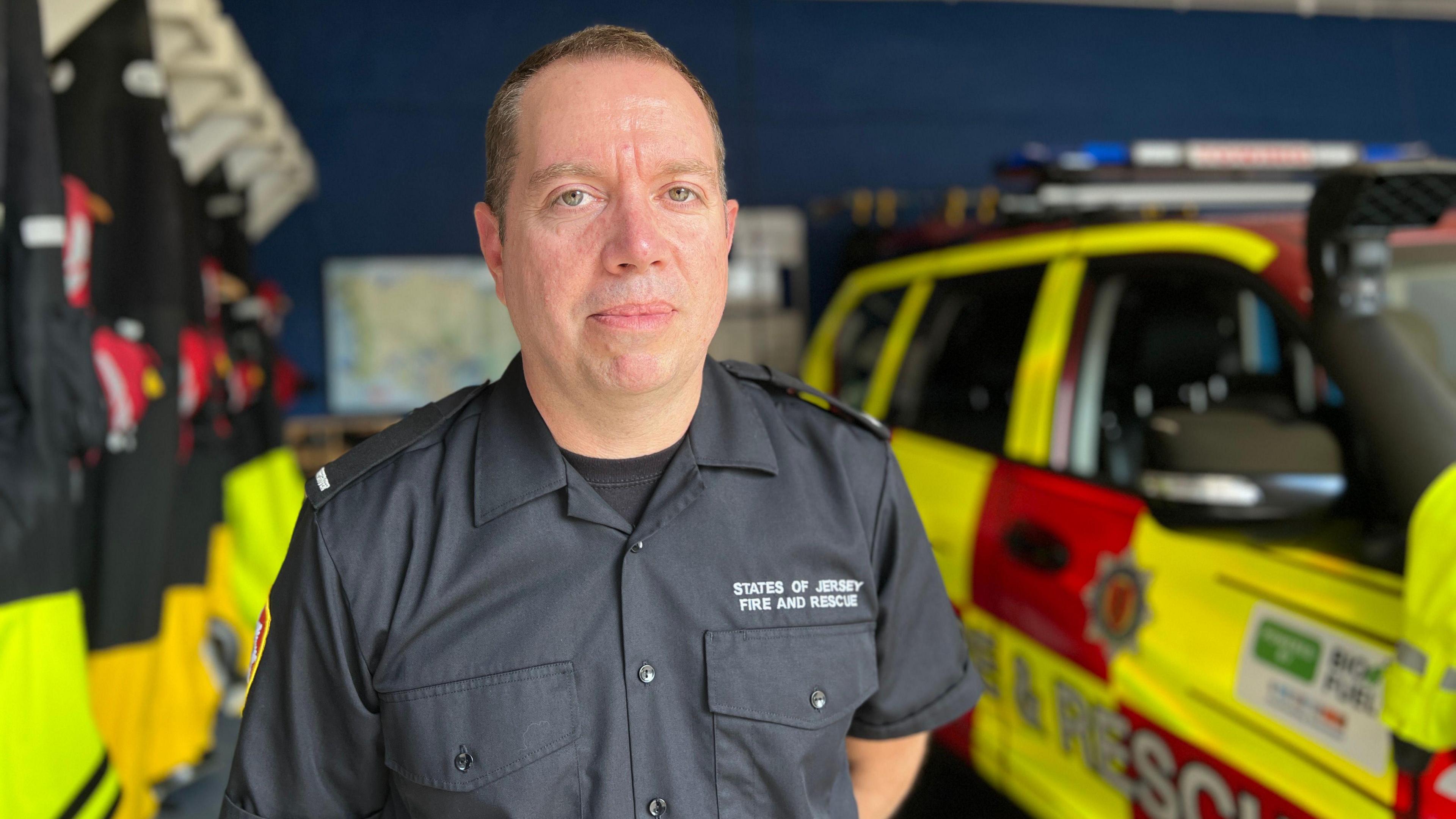 Firefighter Jonny Burch looks at the camera. He is wearing uniform and has short dark hair and his arms are behind his back. There is a 4x4 fire and rescue vehicle behind him to his right and swimming rescue suits are hanging behind him to his left.