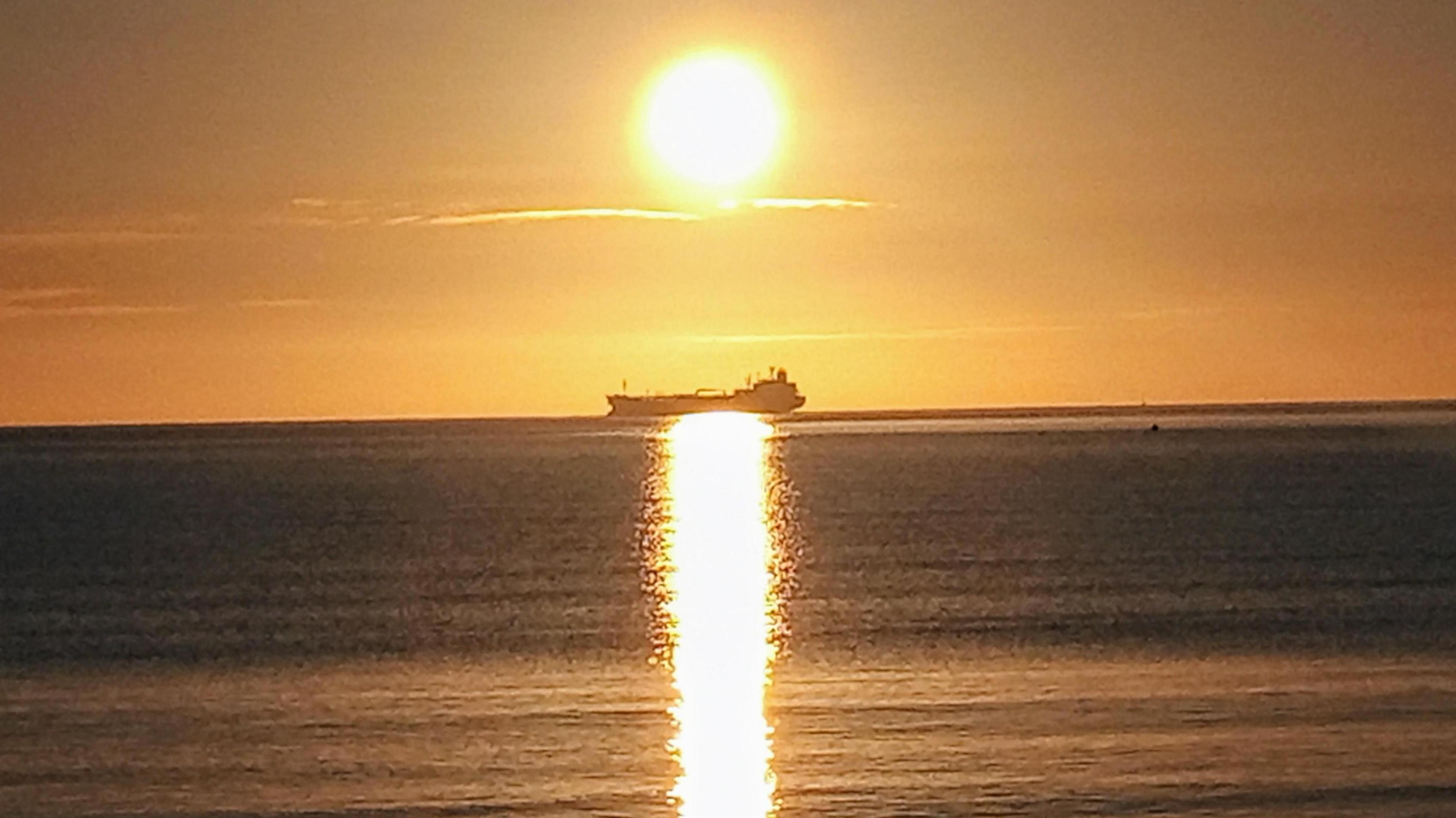 The silhouette of a tanker sails from right to left. It sits perfectly in line with the setting sun and it's reflection on the calm seas. The clear skies are yellow as the sun heads towards the horizon.