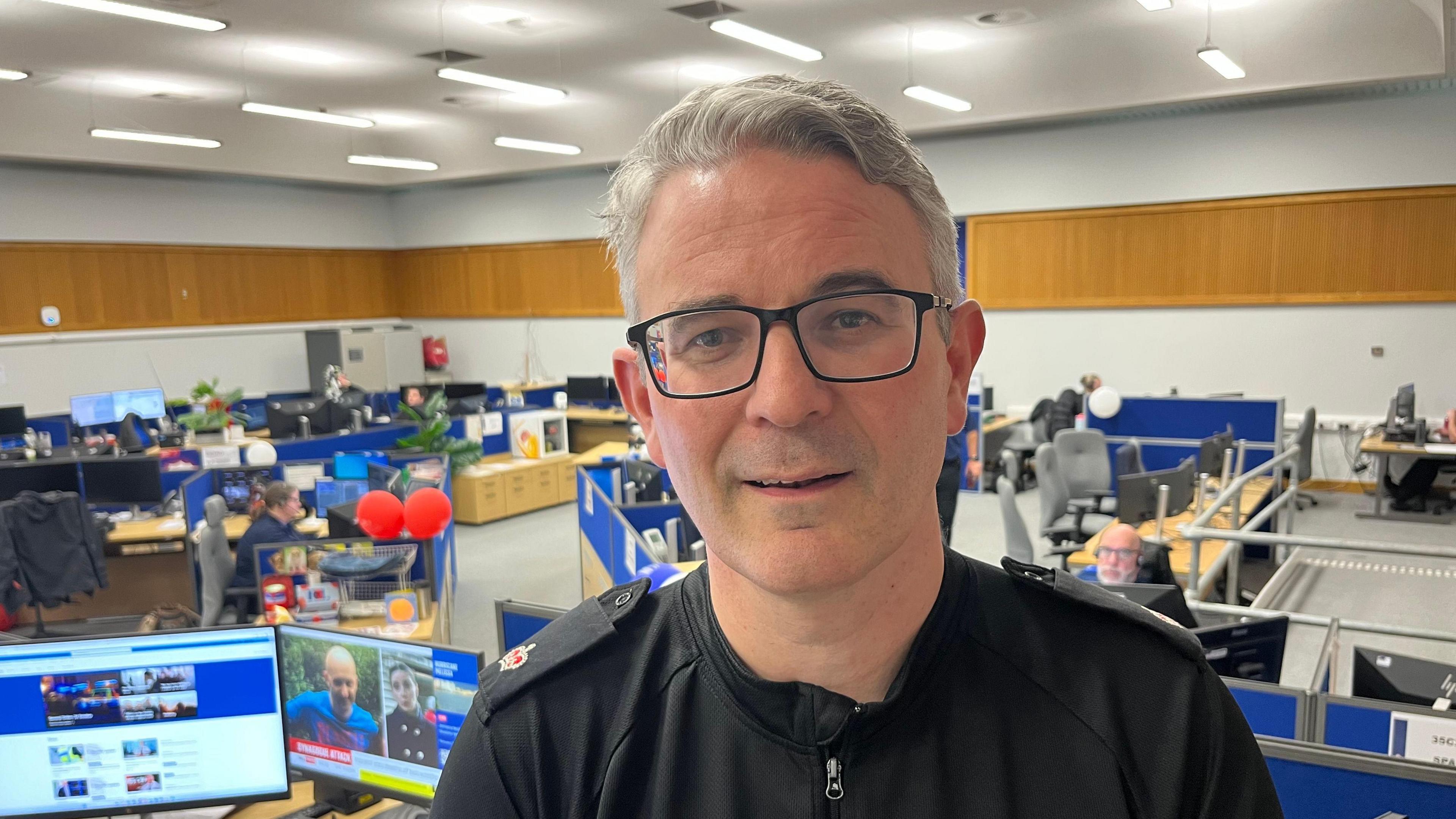 Supt Nick Church is standing on a landing above a control room. He has short, grey hair and is wearing spectacles and a black top with police epaulettes. Behind him, people can be seen sitting at desks with monitors and TV screens on them