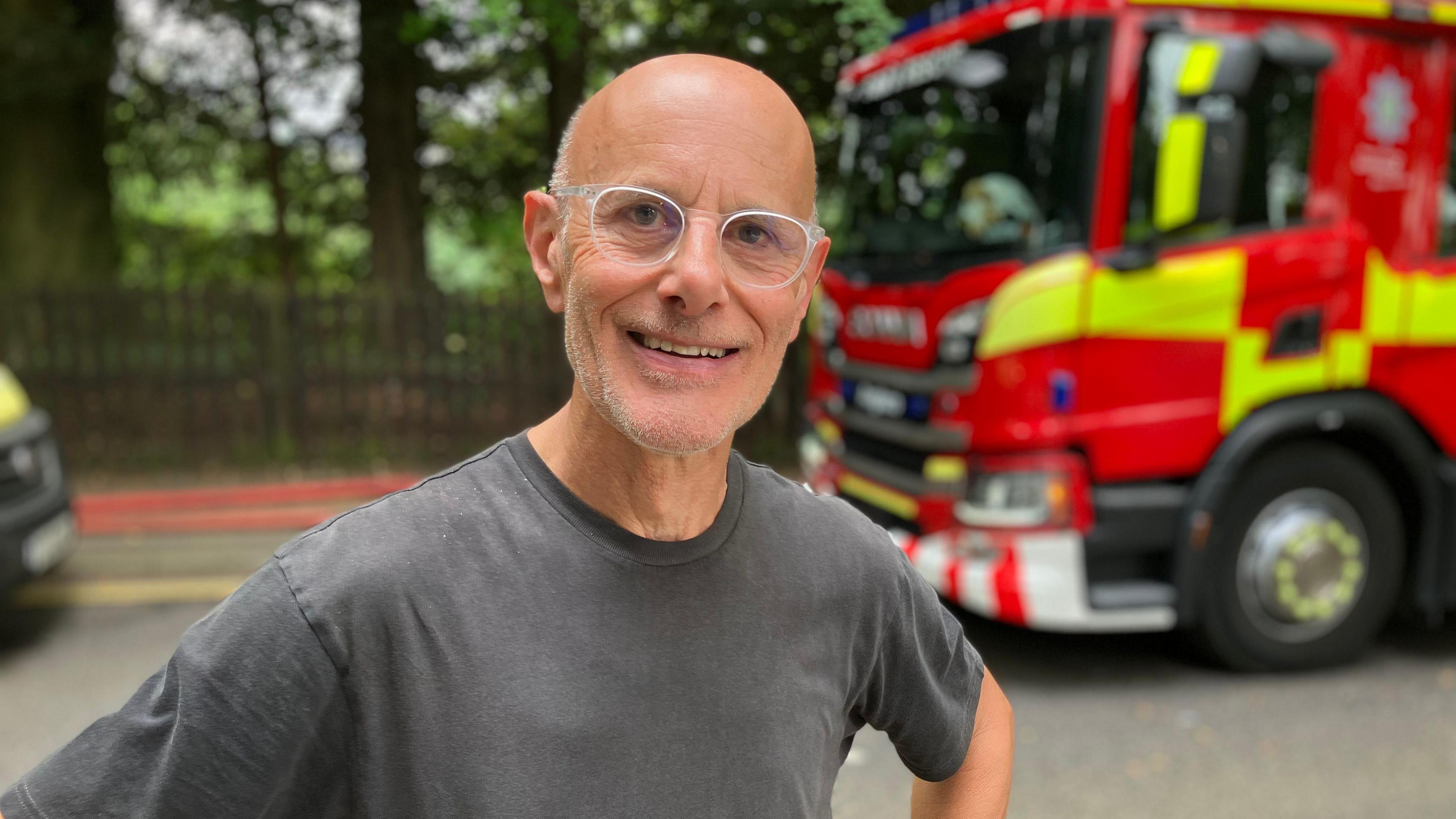 A man with white-rimmed glasses smiling into the camera. He is bald and has a grey top on. In the background is a fire engine with a fence and trees next to it.
