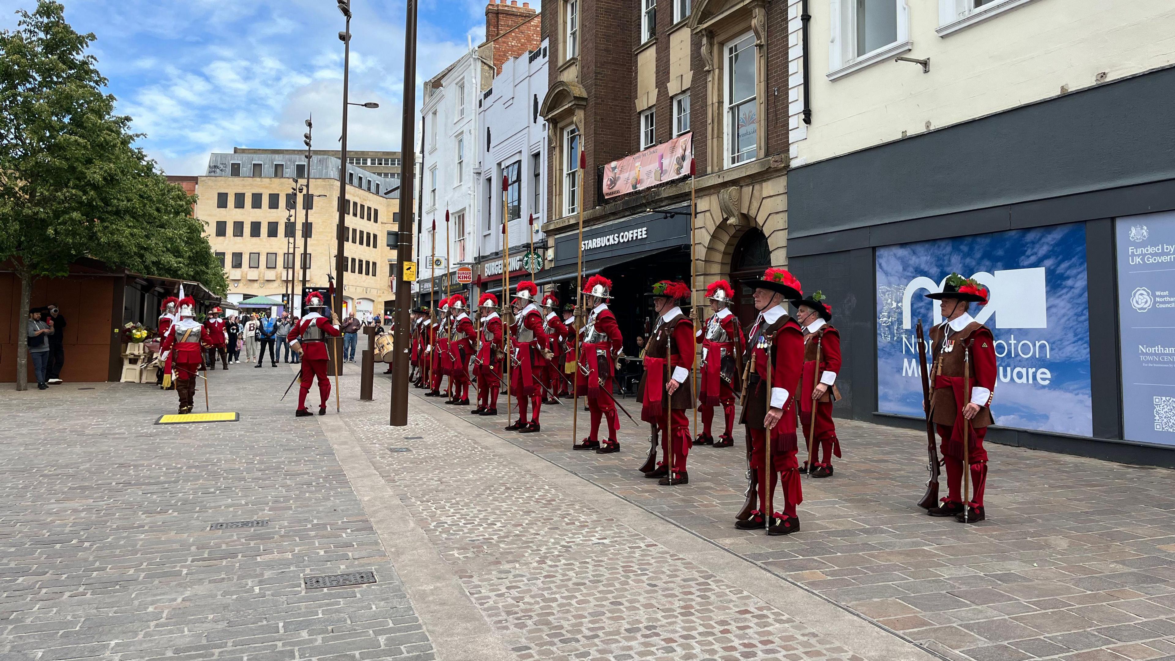 Lots of people dressed in red, black and white medieval military uniforms stand to attention in a town centre.