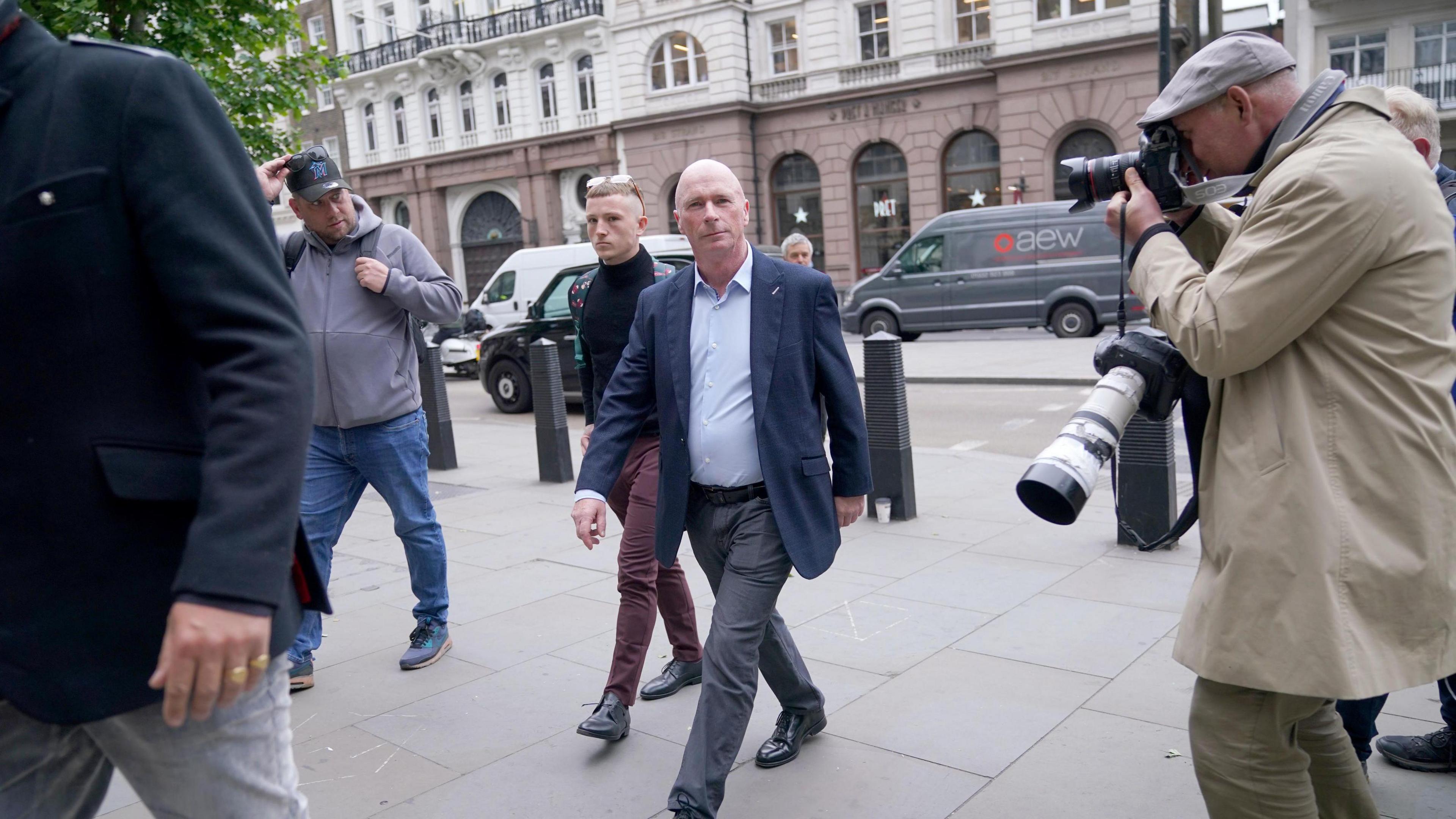 Ray Connolly with very short light-coloured hair, striding across a pavement wearing a blue jacket, light blue shirt and grey trousers. There is a press photographer with a long lens camera on his arm and a smaller camera up to his eye. Other people are visible and there is a grey van parked on the other side of the road.