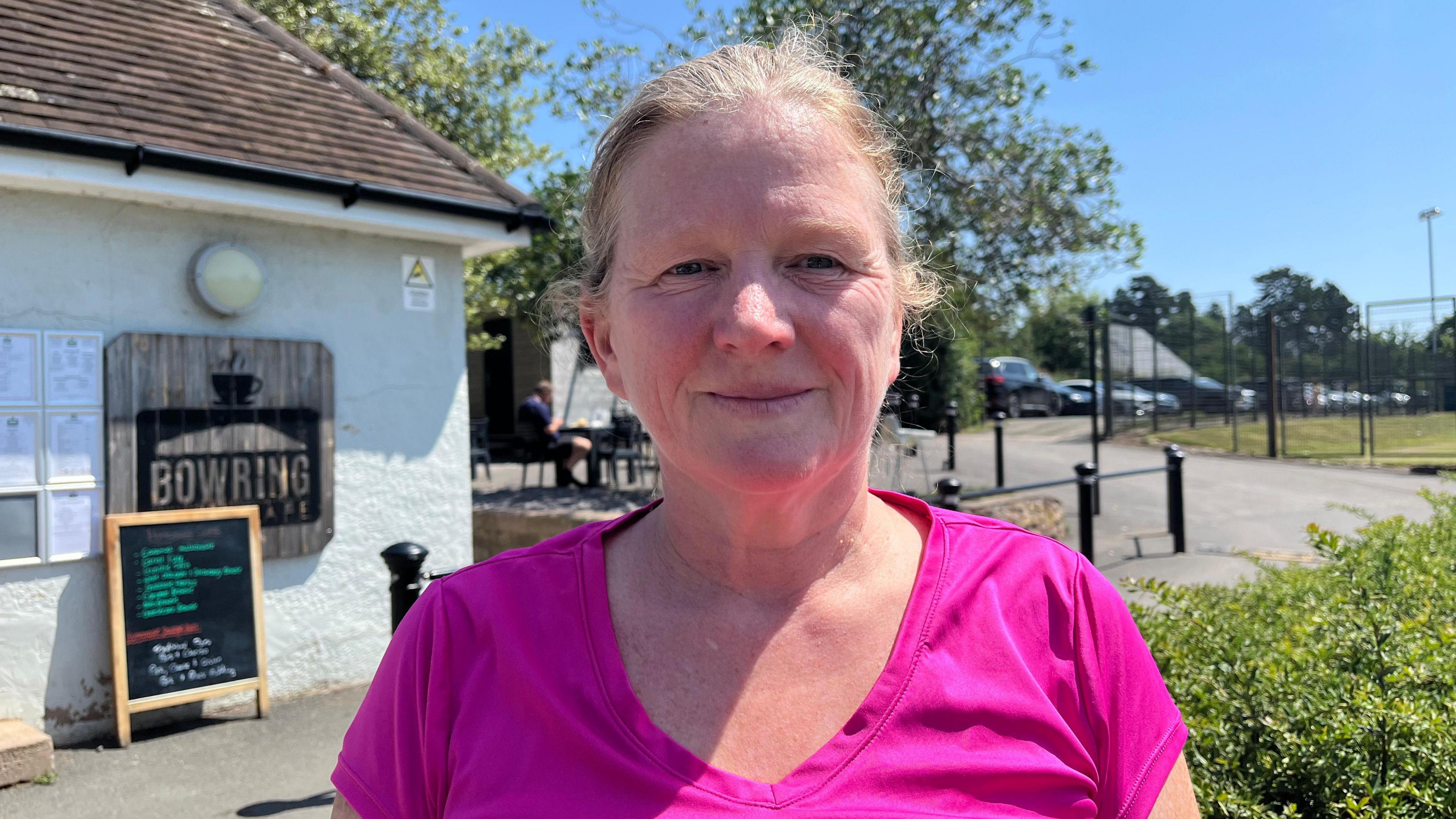 Donna pictured looking into the camera and smiling slightly. She has blonde grey hair tied up and is wearing a hot pink athletic top. She is standing outside the Bowring Park Cafe. It is a single-storey, small building, painted white and a chalkboard sign is standing outside it.