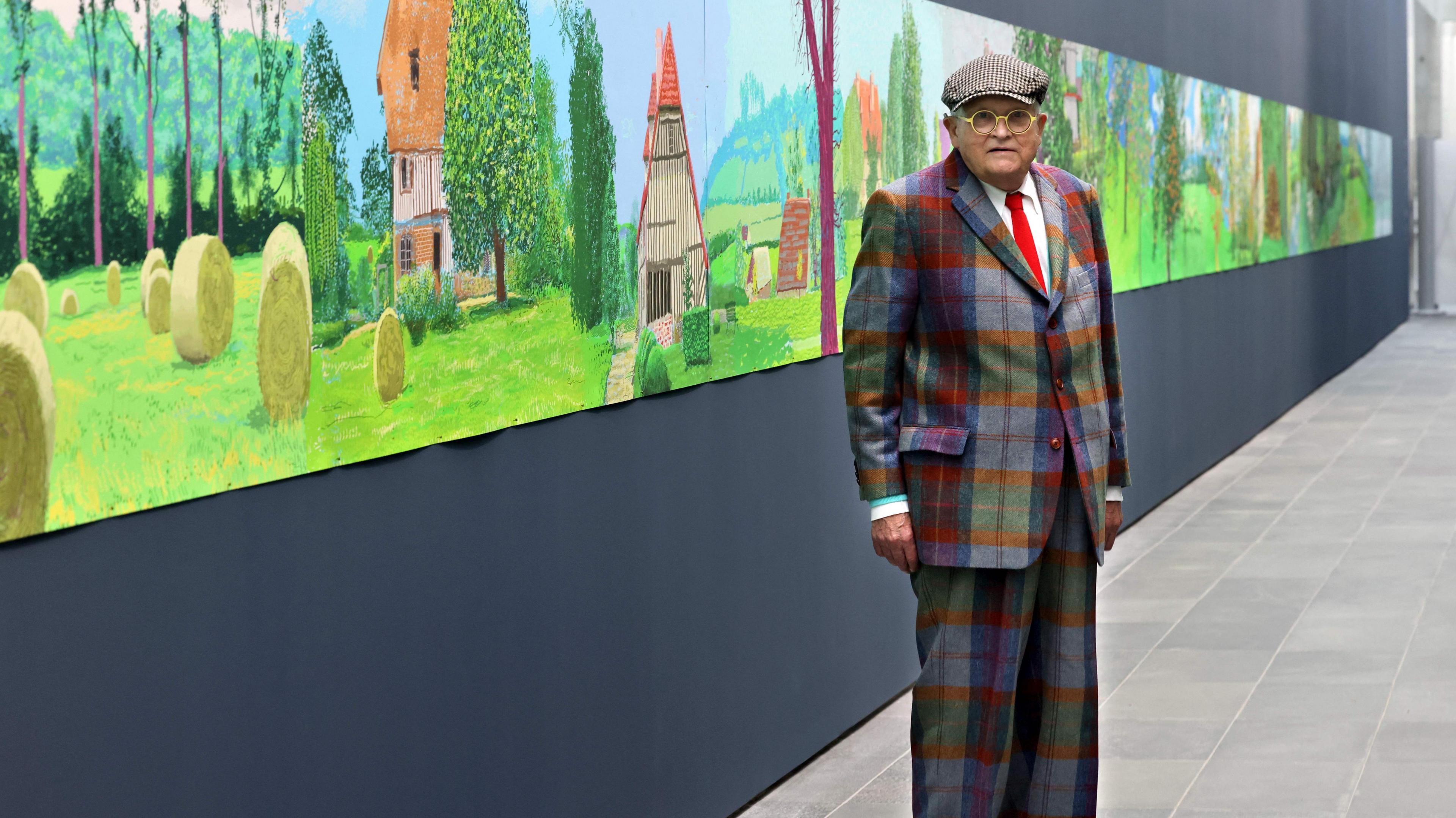 David Hockney in checked suit and hat posing in front of his painting "A year in Normandy", a colourful work showing landscape of Normandy in seasons
