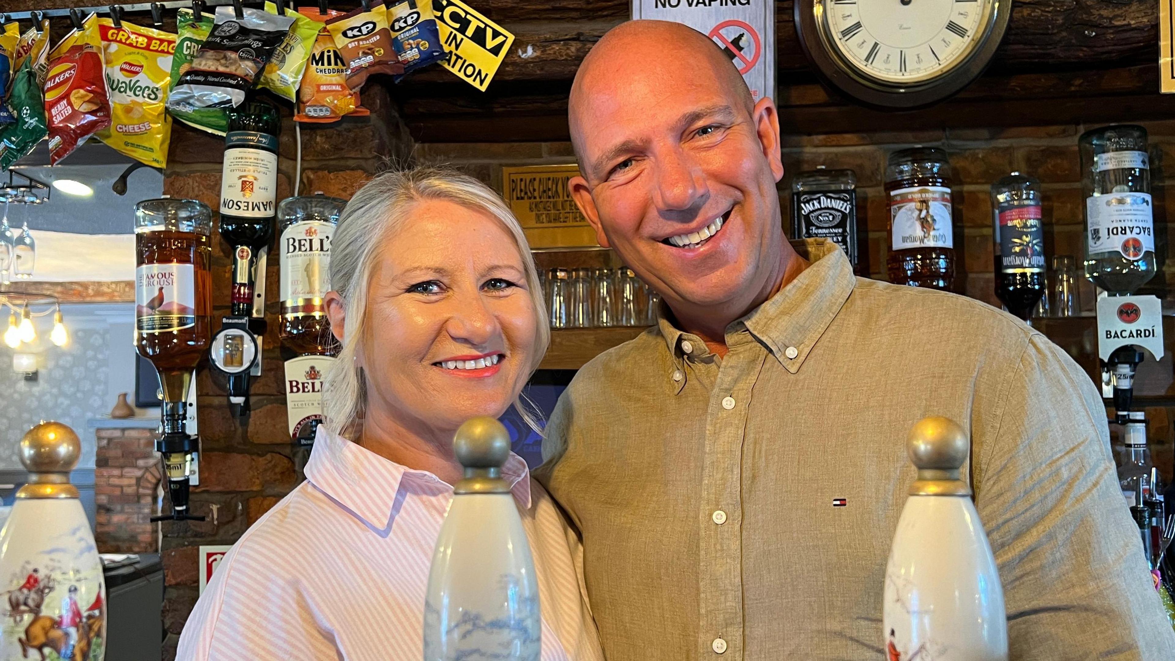 Julie and Lee Loach have happy smiley looks on their faces as they stand behind the bar at their pub. Julie is wearing a striped pink blouse and has blonde hair. Lee is wearing a paisley brown shirt. Behind them, spirit bottles are fixed to the wall and crisp packets are strung above them.