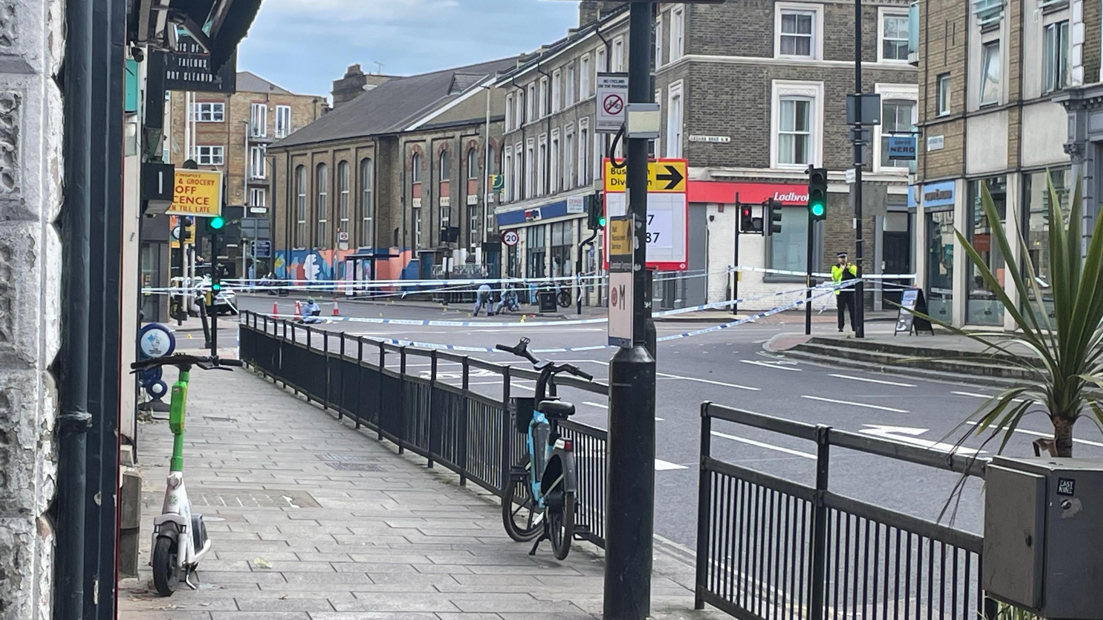 Lavender Road in Clapham at the junction with Wandswoth Road. In the background, forensic officers are seen while police tape is around them. In the foreground is a lamp post, black railings and the pavement and road. 