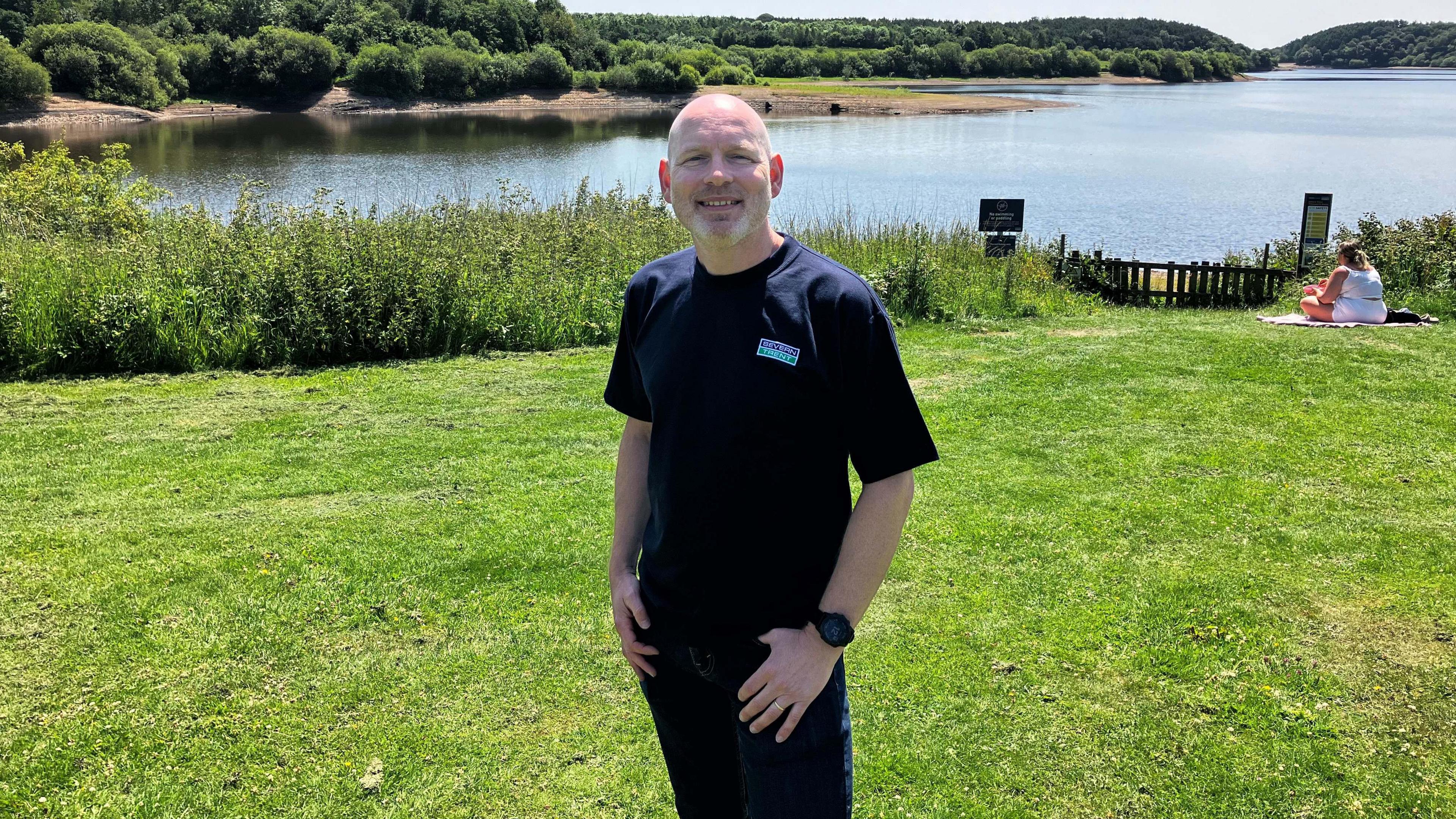 A man wearing a blue T-shirt and jeans stands on grass. There is a reservoir behind him with trees visible on the other side. A woman wearing all white can be seen sitting on the grass behind him to his right.