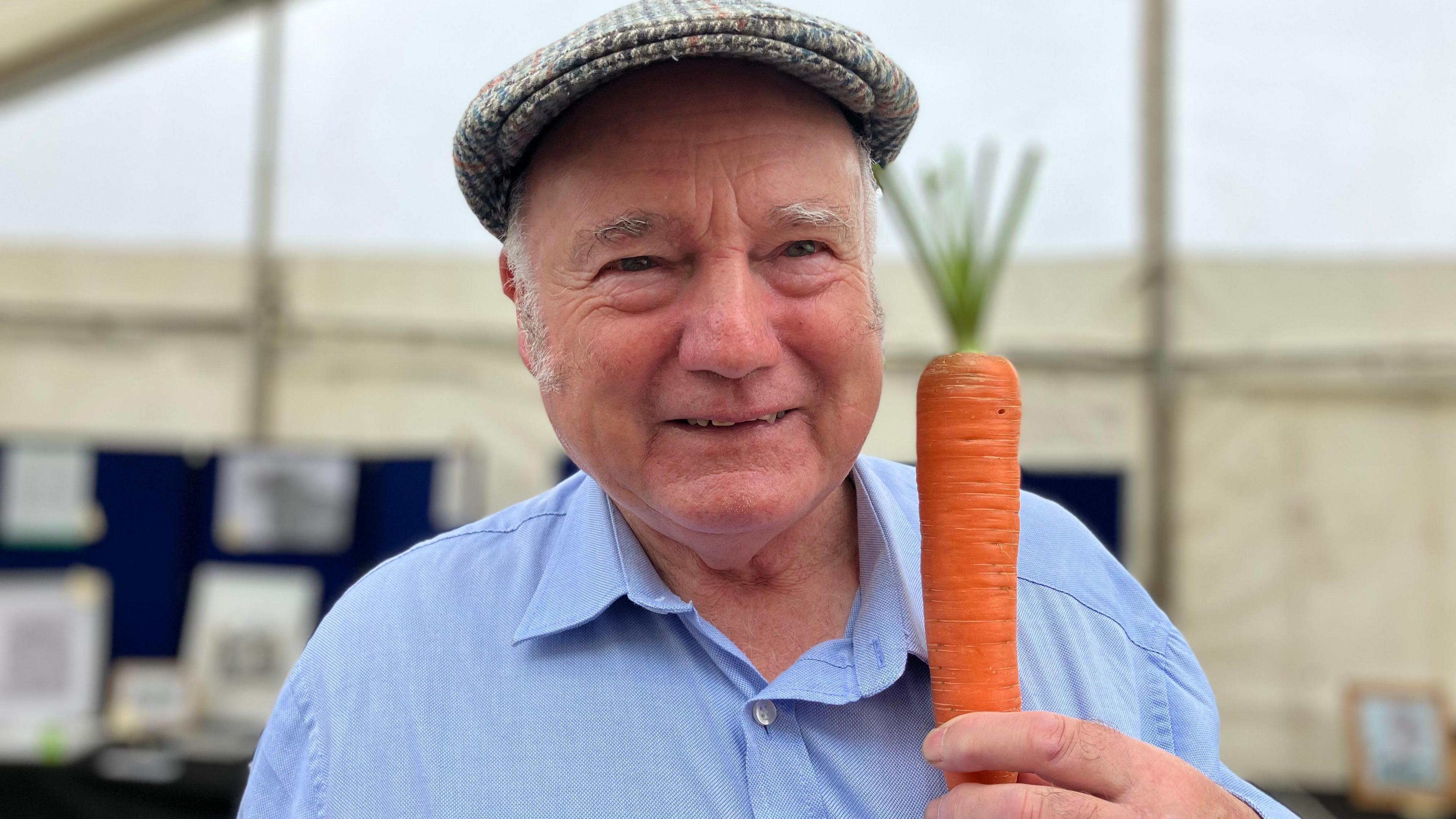 A smiling old man in a peaked cap holds up a carrot.