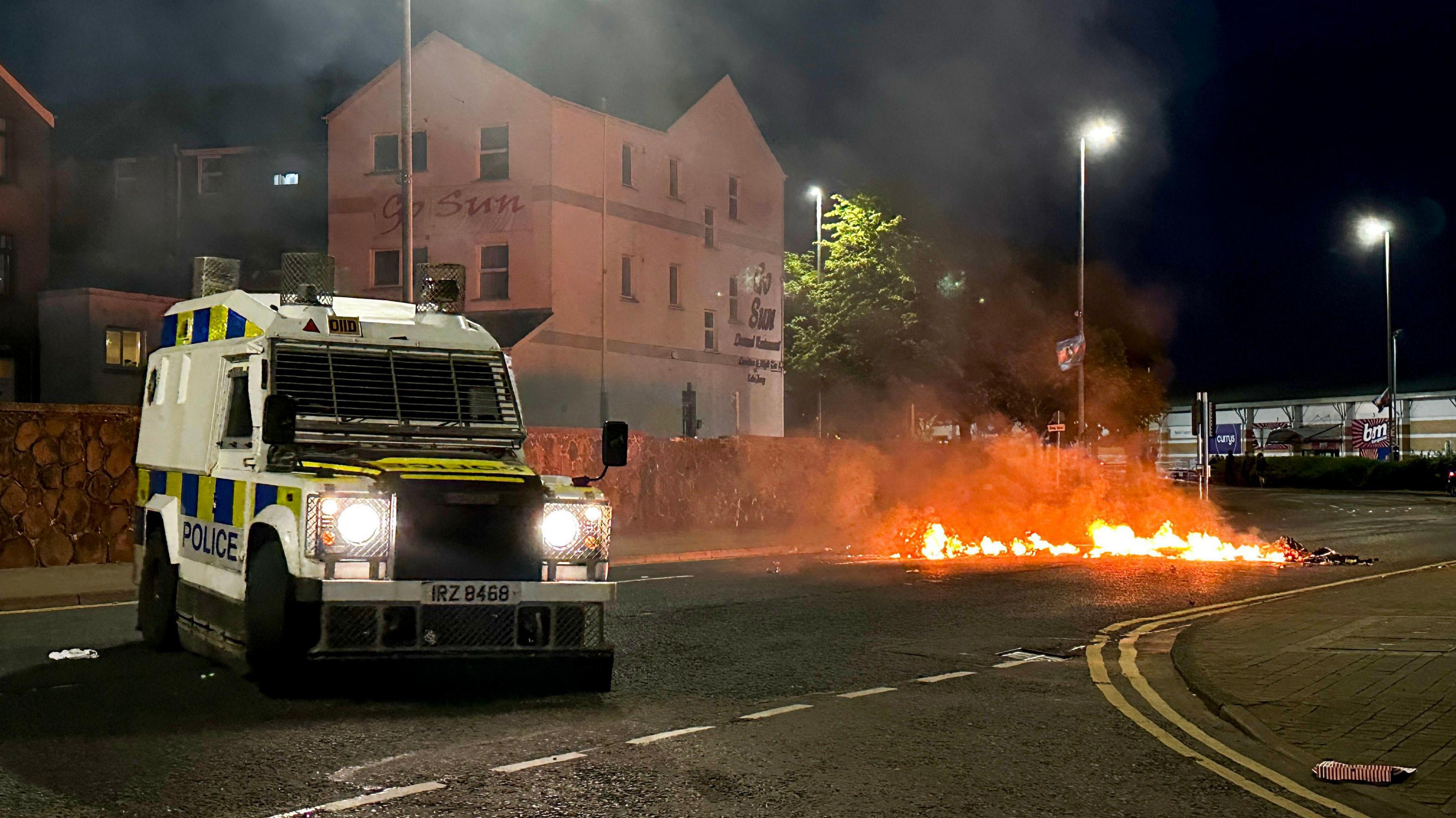 A white police land rover with yellow and blue signage saying POLICE, parked in the middle of the road where a piece of material has been set a light across the road.